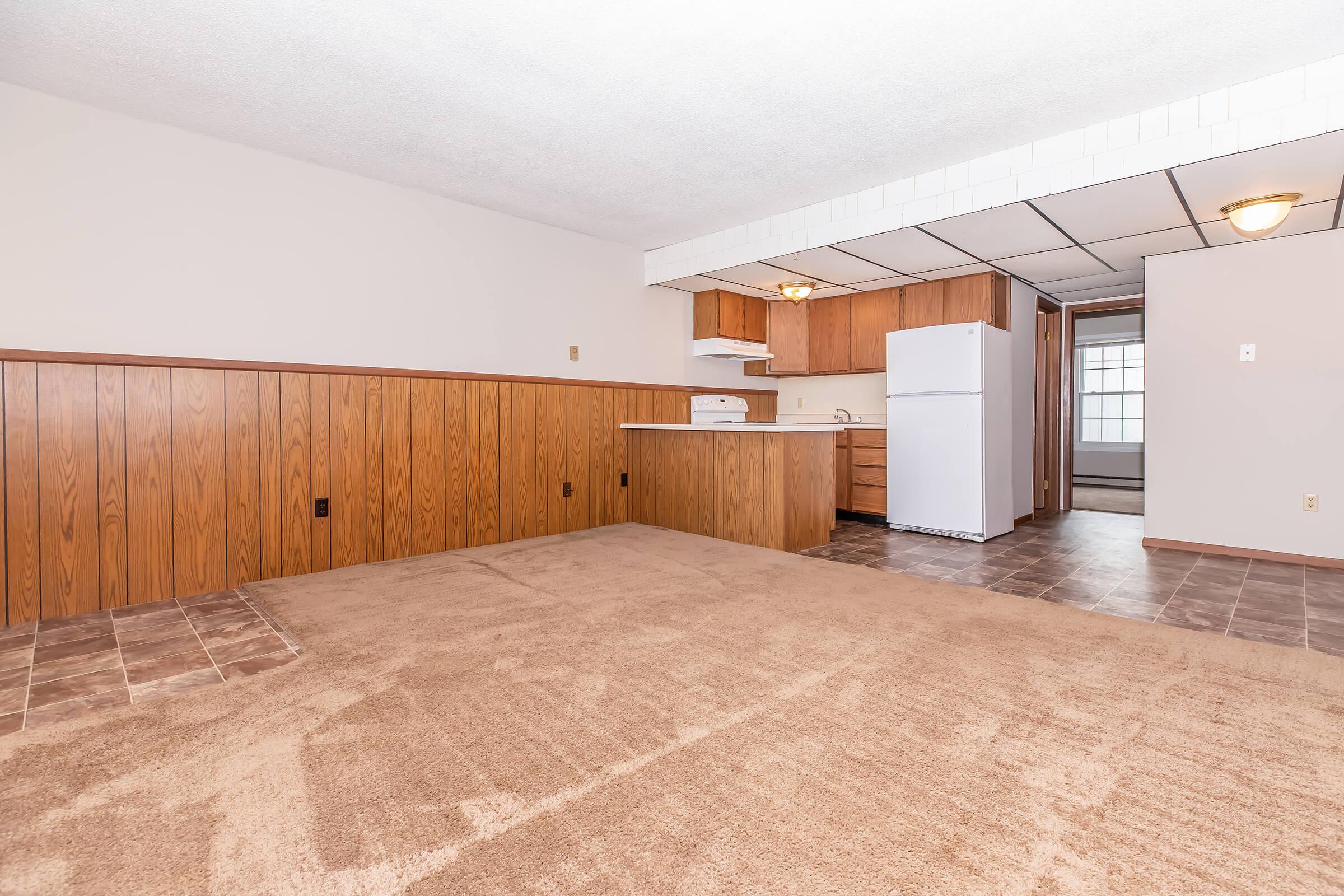 A spacious living area with light beige carpet, wooden paneled walls, and a kitchen in the background. The kitchen features wooden cabinets, a white refrigerator, a stove, and a small island. Natural light comes in from a window at the far end of the room. The space is well-lit and inviting.