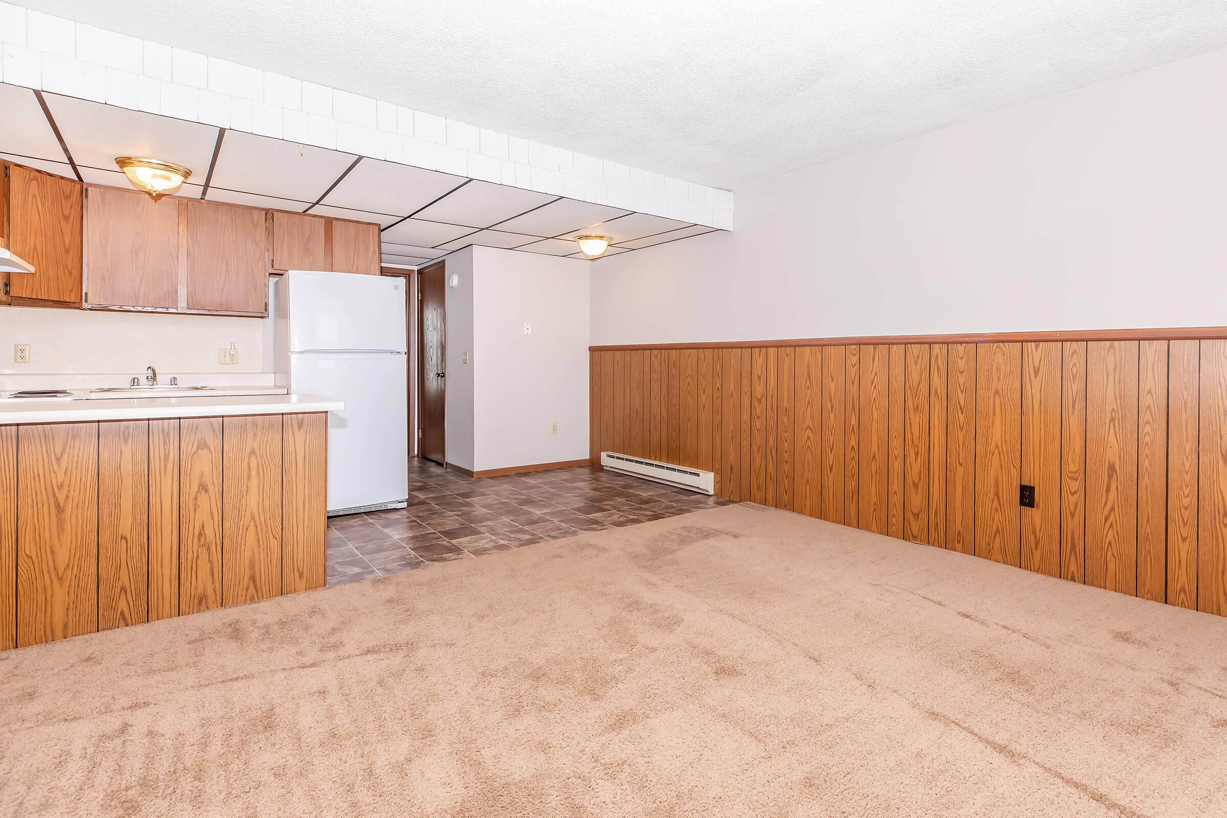 An empty room featuring carpeted flooring and wooden paneling on the lower half of the walls. On one side, there is a small kitchenette with brown cabinets and a white refrigerator. A hallway leads to a door off in the distance, and the space is well-lit by overhead lighting.