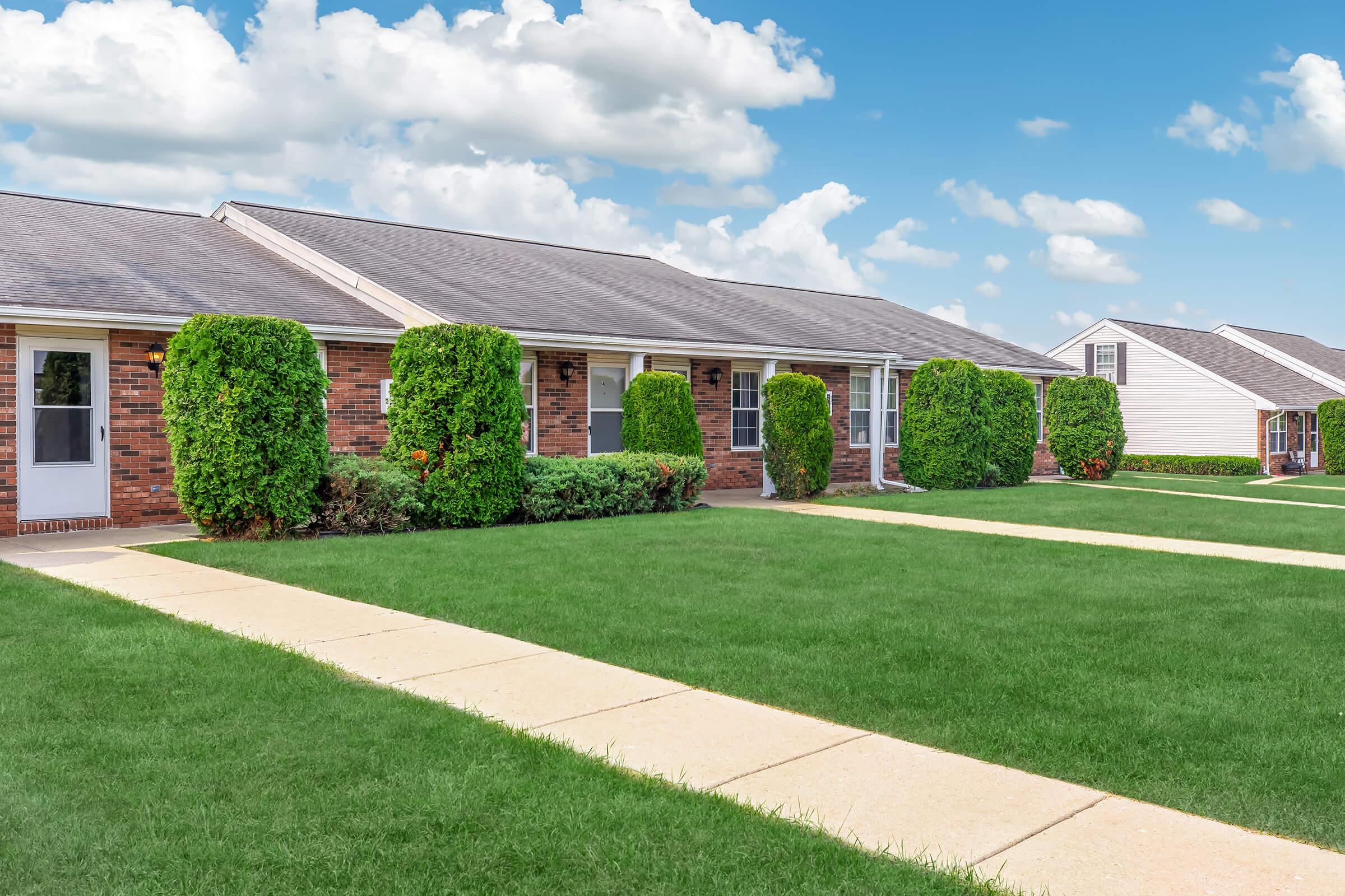 A row of brick duplexes with well-maintained lawns and neatly trimmed hedges. The sky is blue with fluffy white clouds, and concrete walkways lead to the entrances of the apartments. The scene is bright and inviting, showcasing a well-kept residential area.