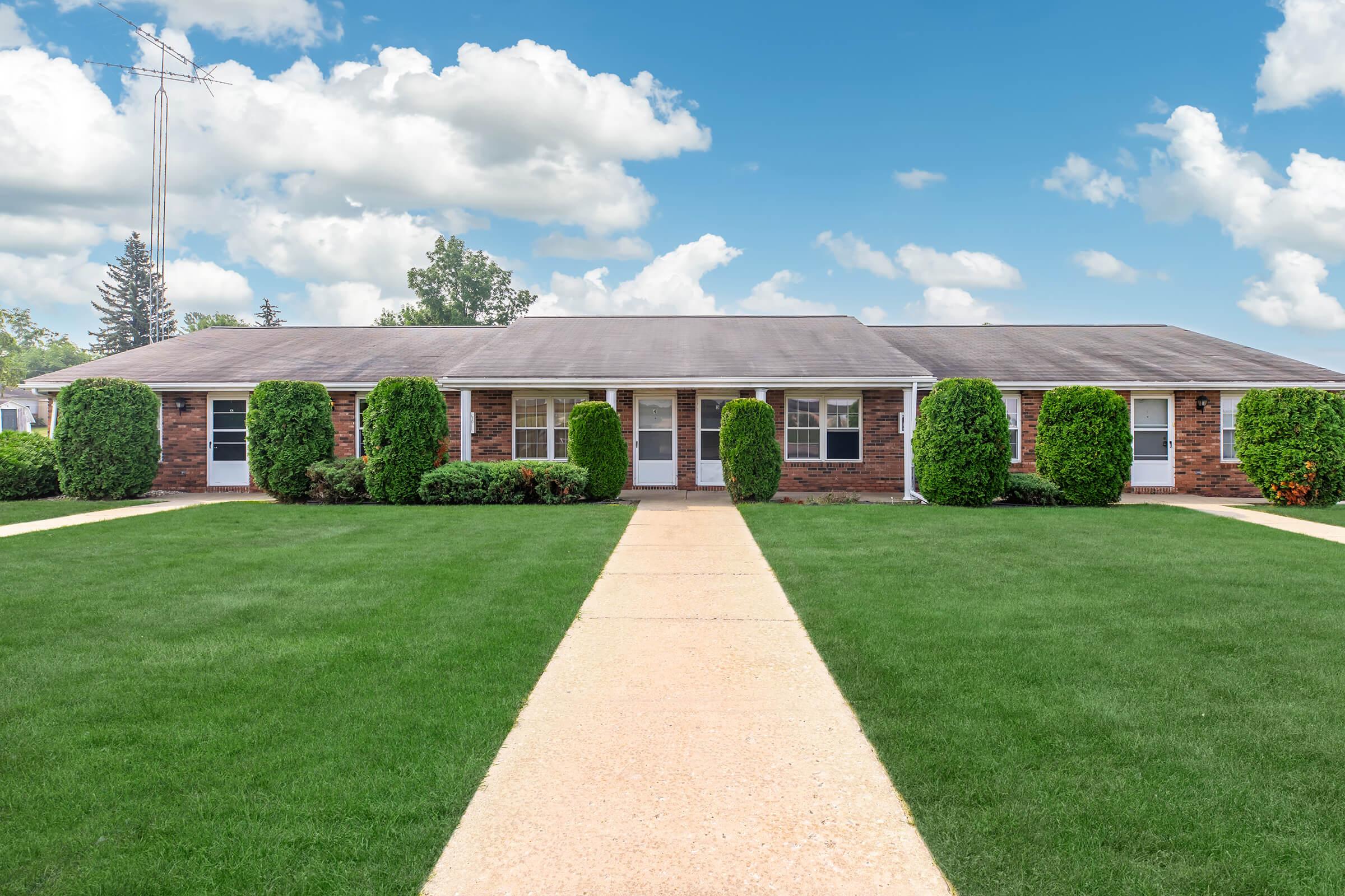 A brick residential building with multiple entrances, surrounded by well-manicured lawns and shrubs. The sky is partly cloudy, adding a bright contrast to the greenery and architecture. A wide concrete pathway leads to the doors of the building.