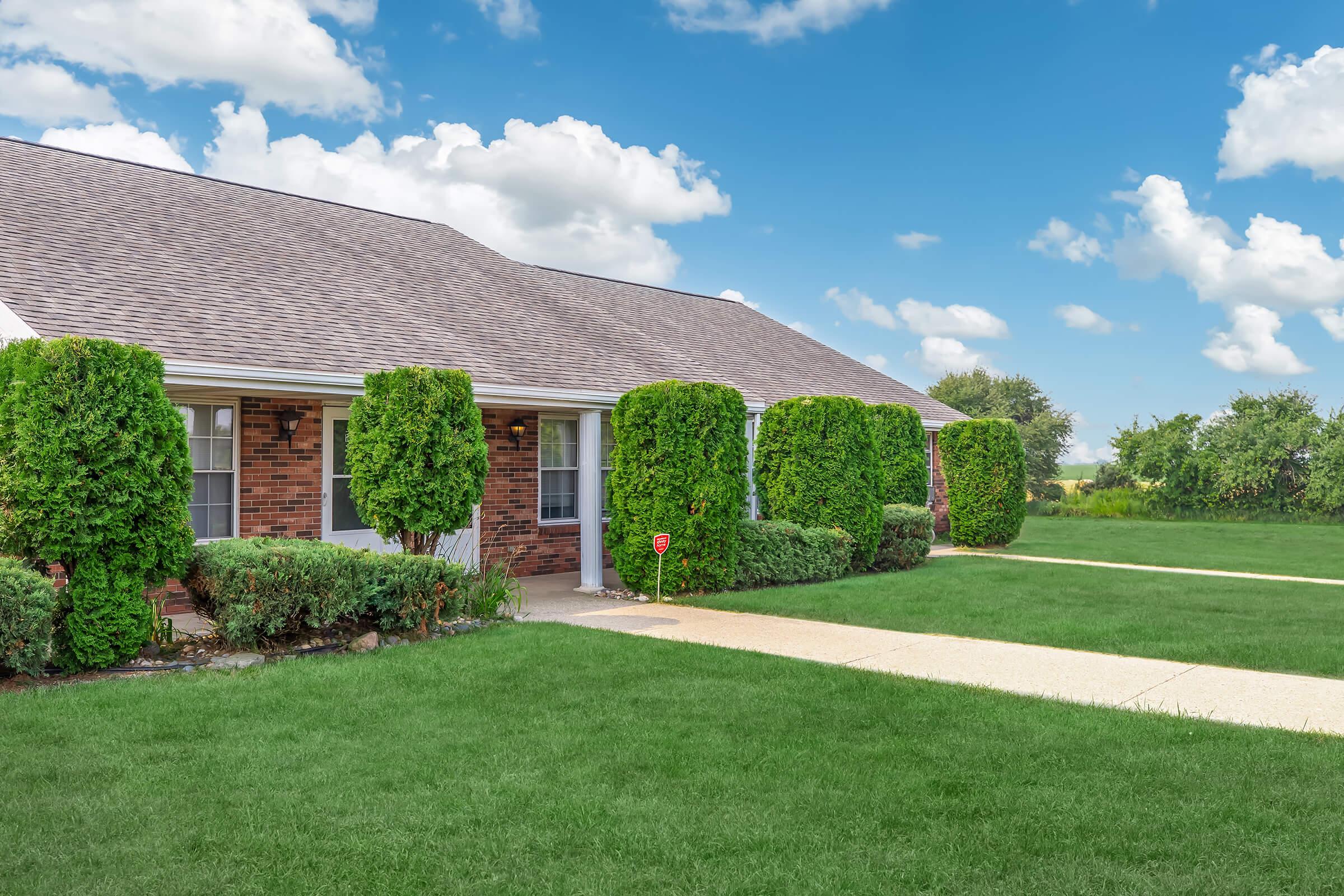 A well-maintained brick house with manicured hedges and a neatly paved walkway, set against a blue sky with fluffy white clouds. Lush green grass surrounds the property, creating a vibrant and inviting outdoor space.