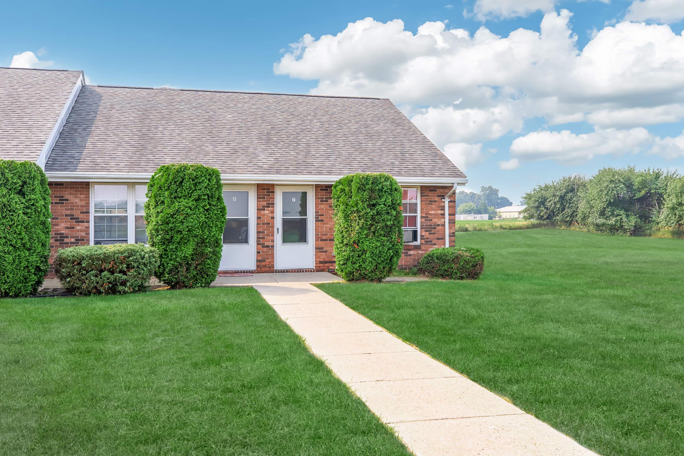 A brick house with a sloped roof features two front doors and well-maintained shrubs on either side. A concrete pathway leads to the entrance, surrounded by lush green grass. In the background, a clear blue sky with fluffy clouds can be seen, adding to the tranquil atmosphere.