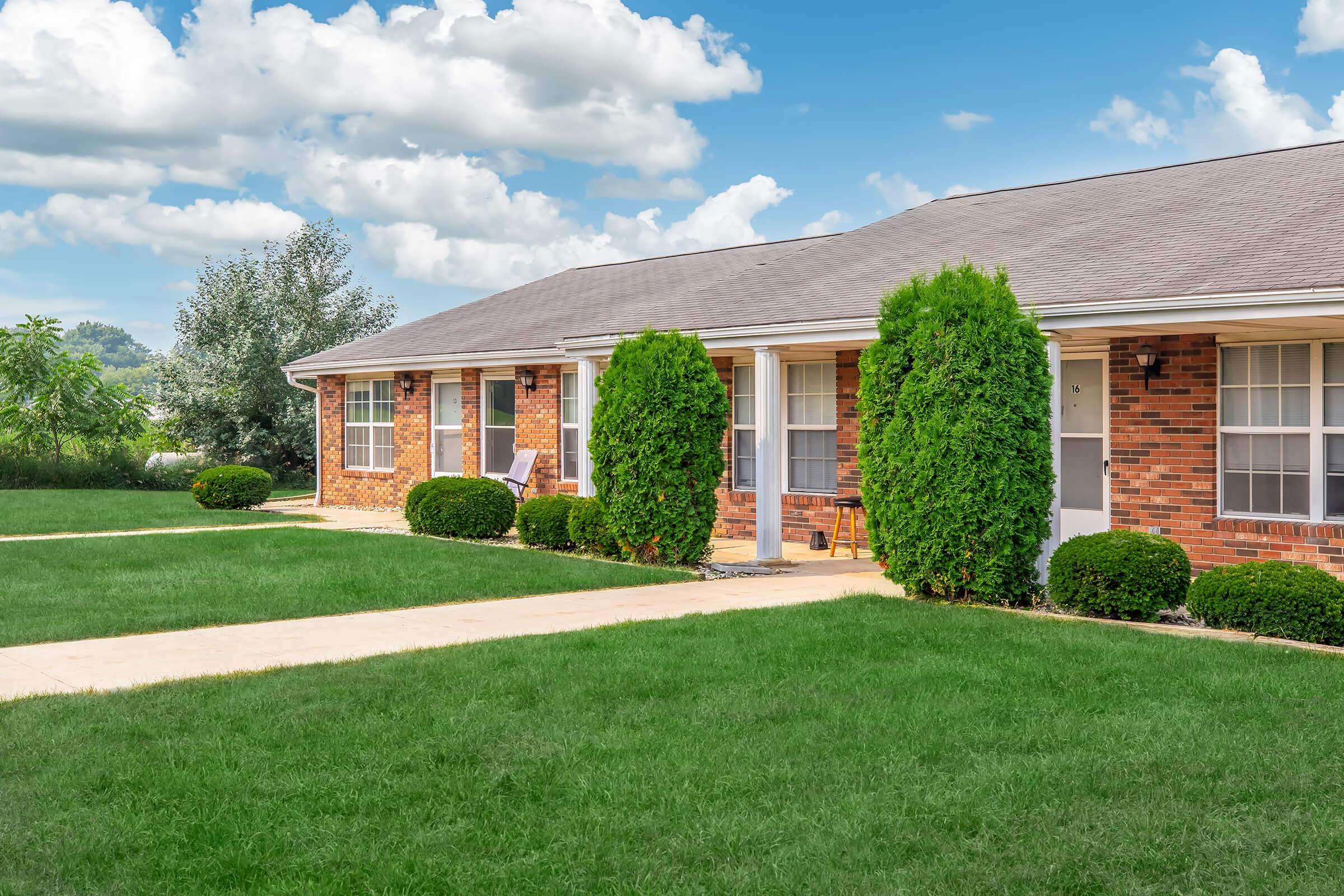 A well-maintained brick apartment building with several units, featuring neat landscaping, green grass, and small shrubs. The sky is partly cloudy, creating a bright and inviting atmosphere. Sidewalks lead to the entrance of each unit, enhancing the inviting appearance of the property.