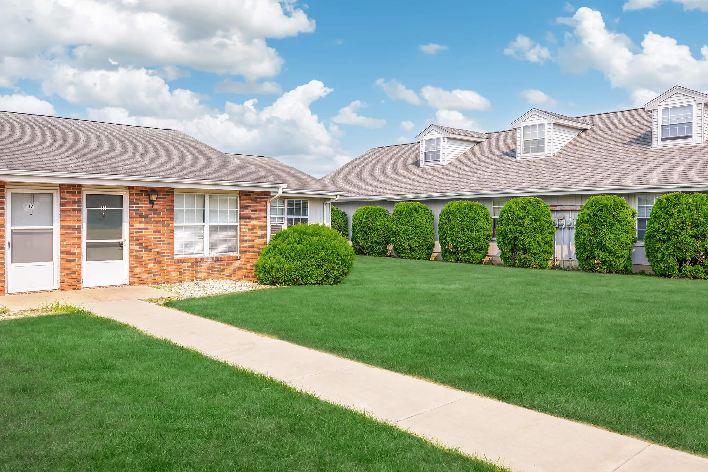 A well-maintained, single-story brick building with a paved walkway leading up to it. The building features multiple windows and a door. Lush green grass surrounds the property, with neatly trimmed shrubbery and another building visible in the background under a bright, cloudy sky.