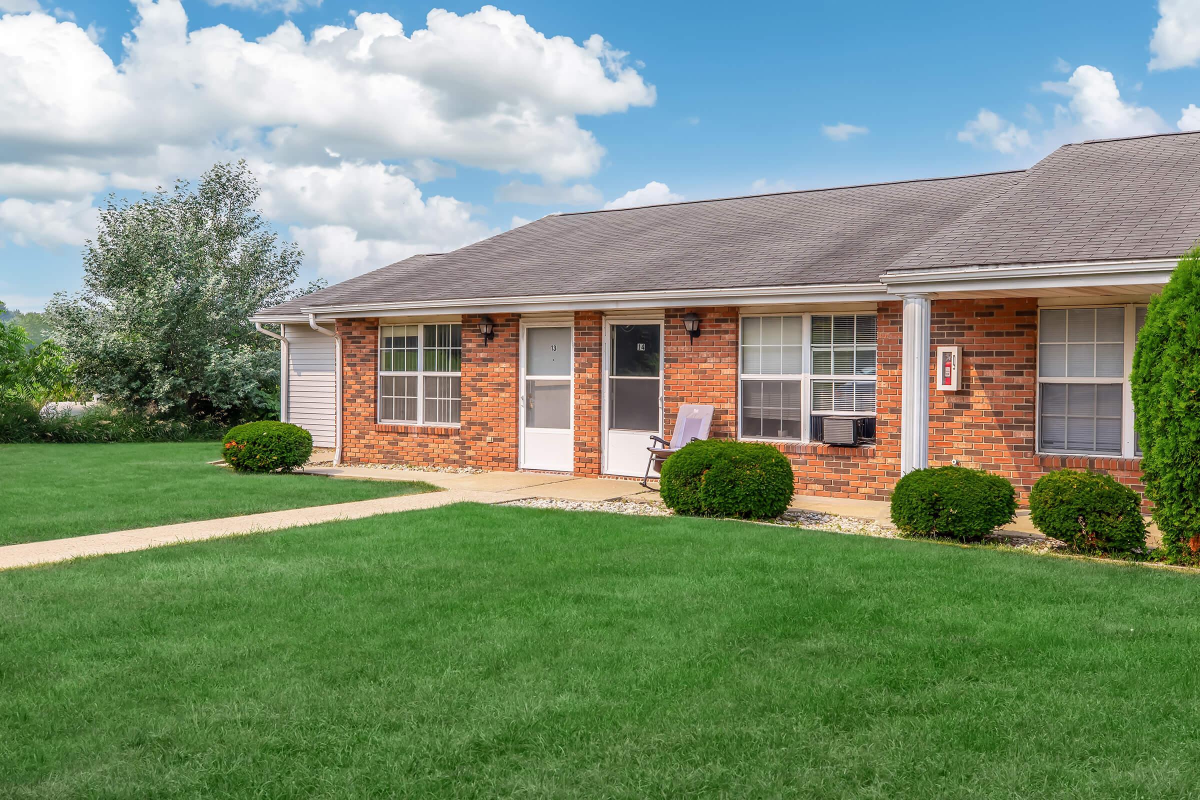 A well-maintained brick apartment building with a grassy lawn, featuring a front porch with a rocking chair, air conditioning unit, and neatly trimmed bushes. The sky is partly cloudy, creating a bright and inviting atmosphere.