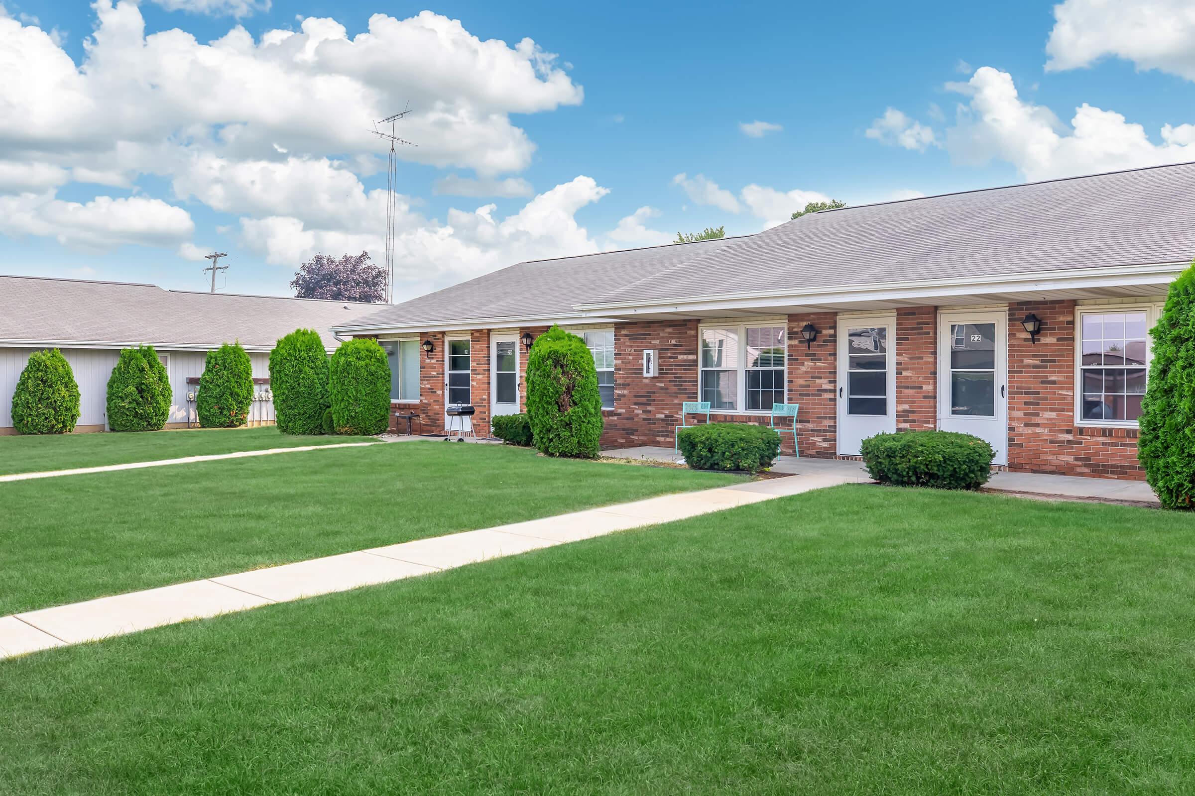 A well-kept single-story brick apartment complex with a neat green lawn. The building features multiple entrance doors with windows beside them, bordered by small shrubs. The sky is clear with fluffy white clouds, creating a bright and inviting atmosphere.