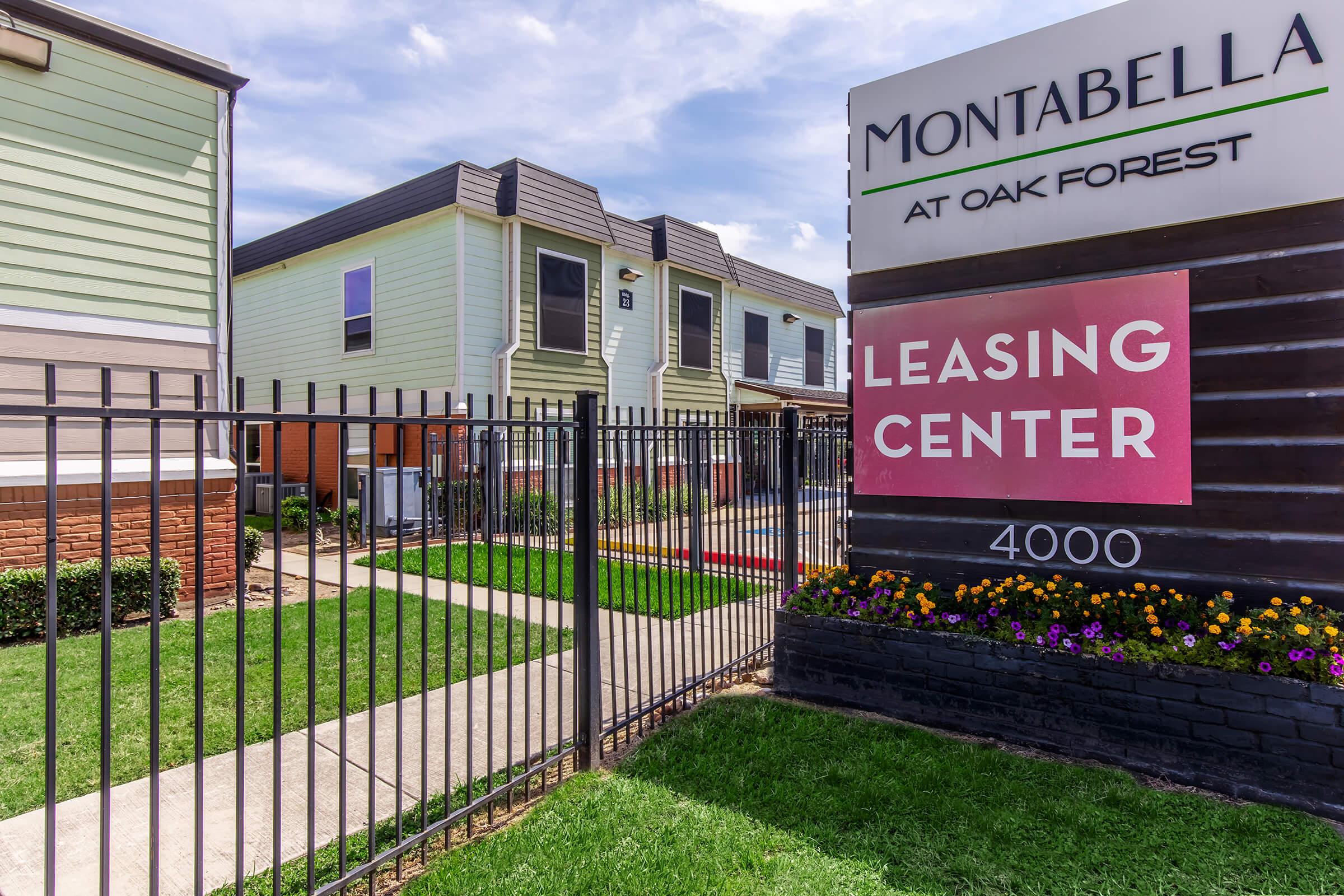 A view of Montabella at Oak Forest, featuring a leasing center sign and a fenced entrance. The buildings are modern, with greenery and colorful flowers in the landscaping, creating an inviting atmosphere. The address visible is 4000.