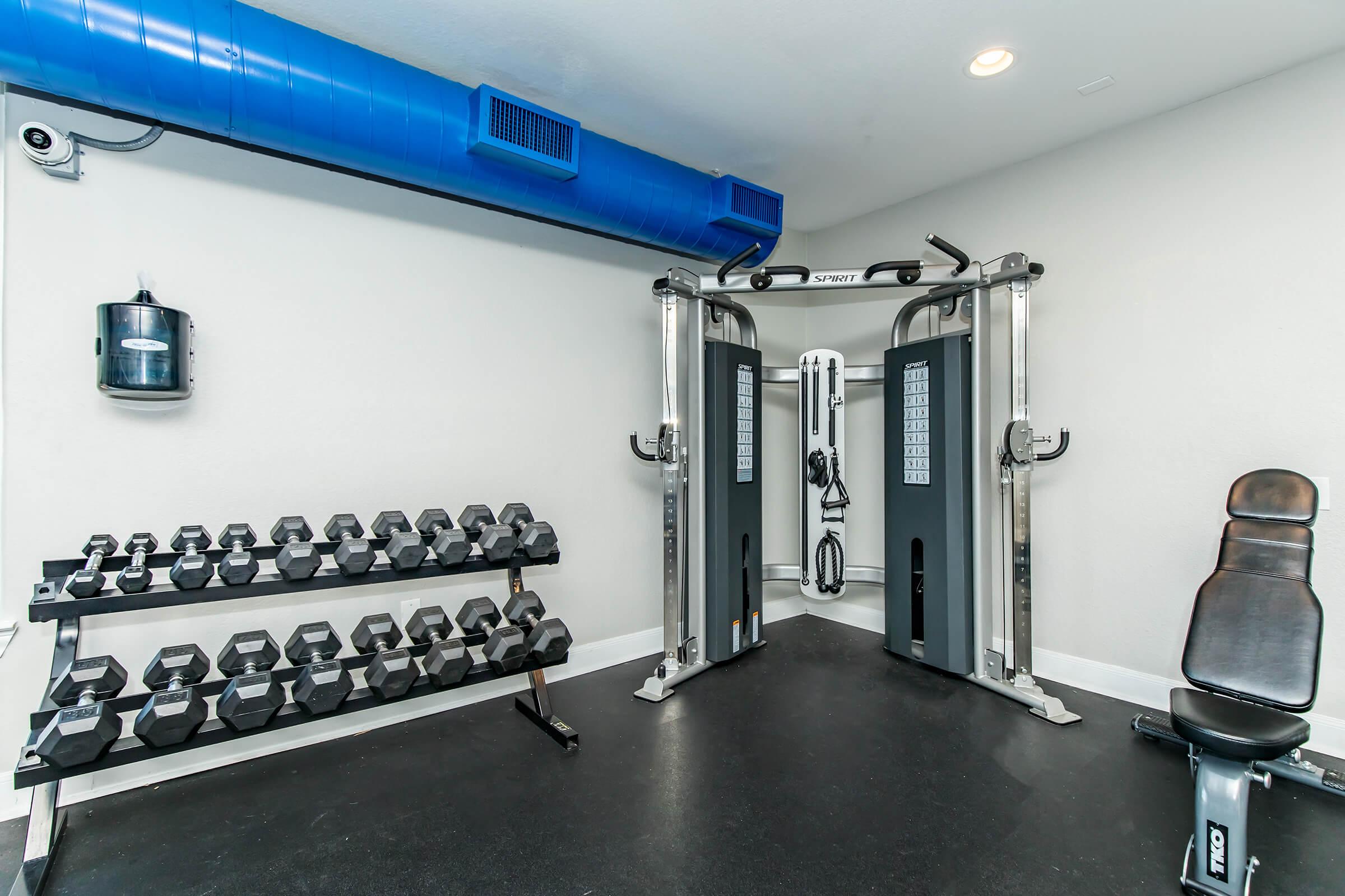 A small home gym setup featuring a cable machine with two towers, a weight bench, and a rack of dumbbells on the side. The room has a clean, modern look with light-colored walls and a blue ventilation duct along the ceiling. The floor is black and designed for exercise.