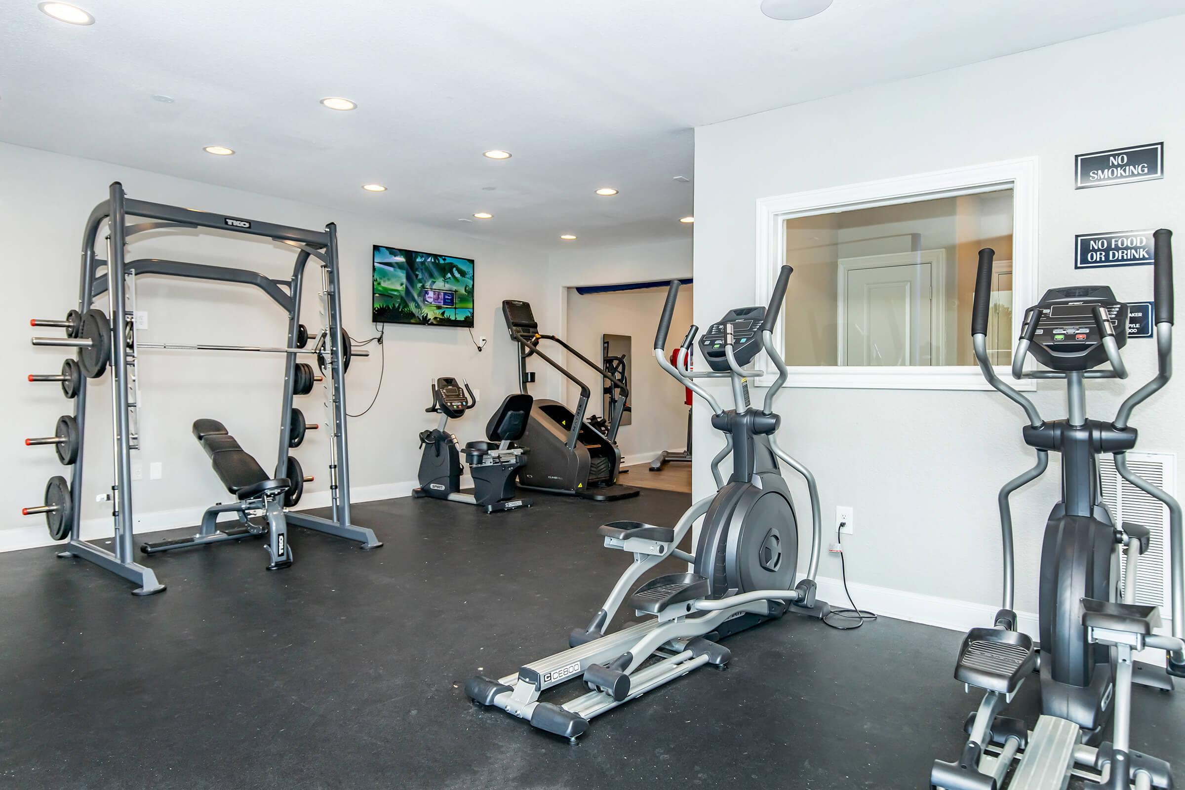 Modern gym interior featuring various exercise equipment including a multi-station weight machine, elliptical, and treadmill. The space has rubber flooring, bright lighting, and a large window with a view. A television is mounted on the wall, and signage indicates "No Smoking" and "No Food or Drink."