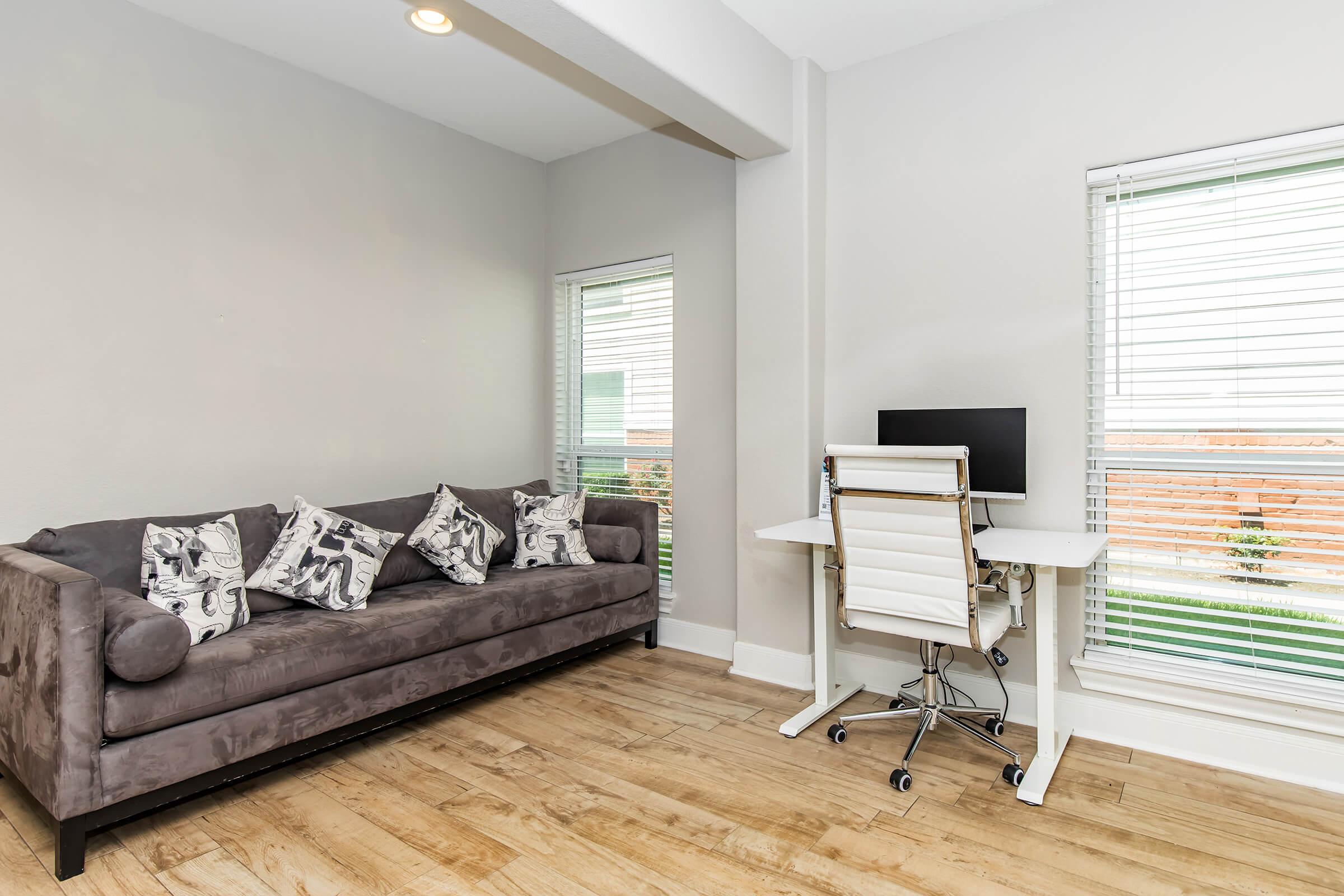 A modern, minimalistic room featuring a gray couch adorned with patterned pillows, a white desk with a computer chair, and large windows with blinds. Soft wooden flooring adds warmth to the space, and the overall design conveys a calm and inviting atmosphere.
