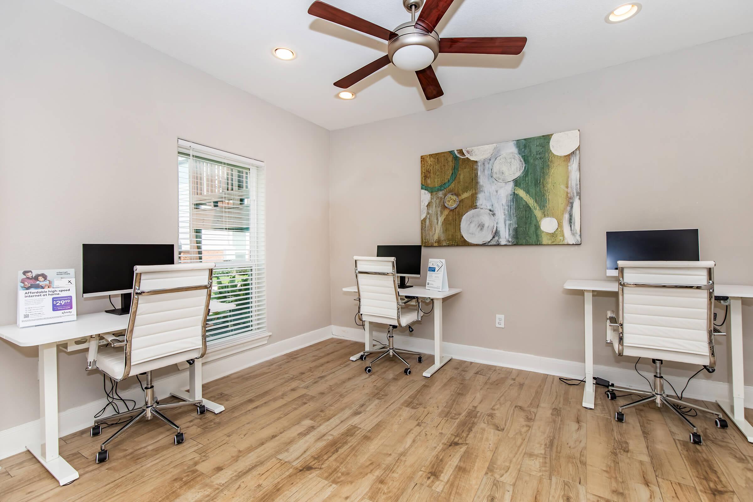A bright home office featuring two modern workstations with computers, white chairs, and a wooden floor. A ceiling fan provides ventilation, and a colorful abstract painting adorns the wall, while natural light streams in through a window.