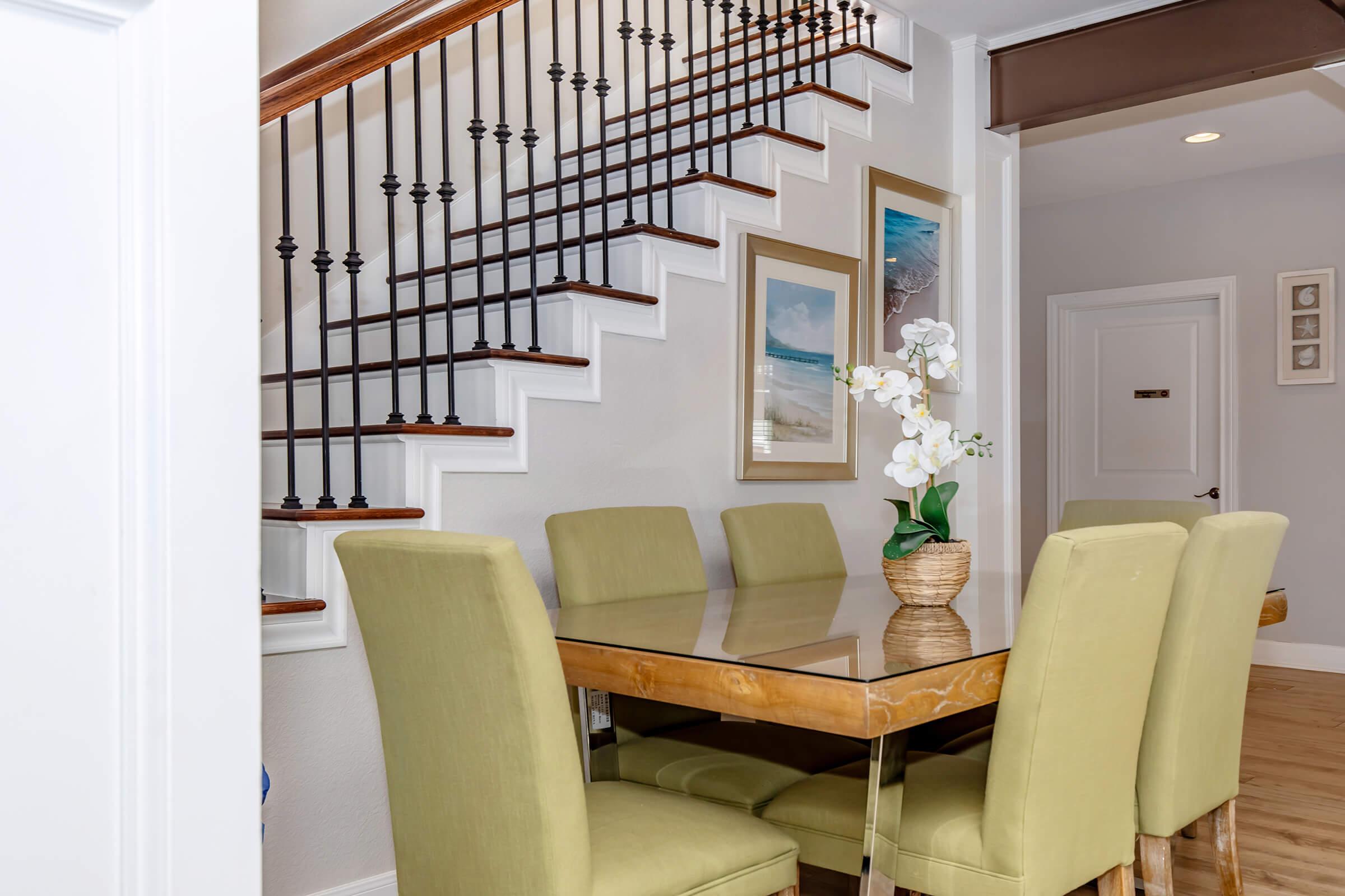 A modern dining area featuring a glossy wooden table surrounded by green upholstered chairs. In the background, a staircase with wooden steps and metal railings ascends. Two framed pictures adorn the wall, and a potted orchid adds a touch of elegance to the table. Light colors create a bright and inviting atmosphere.