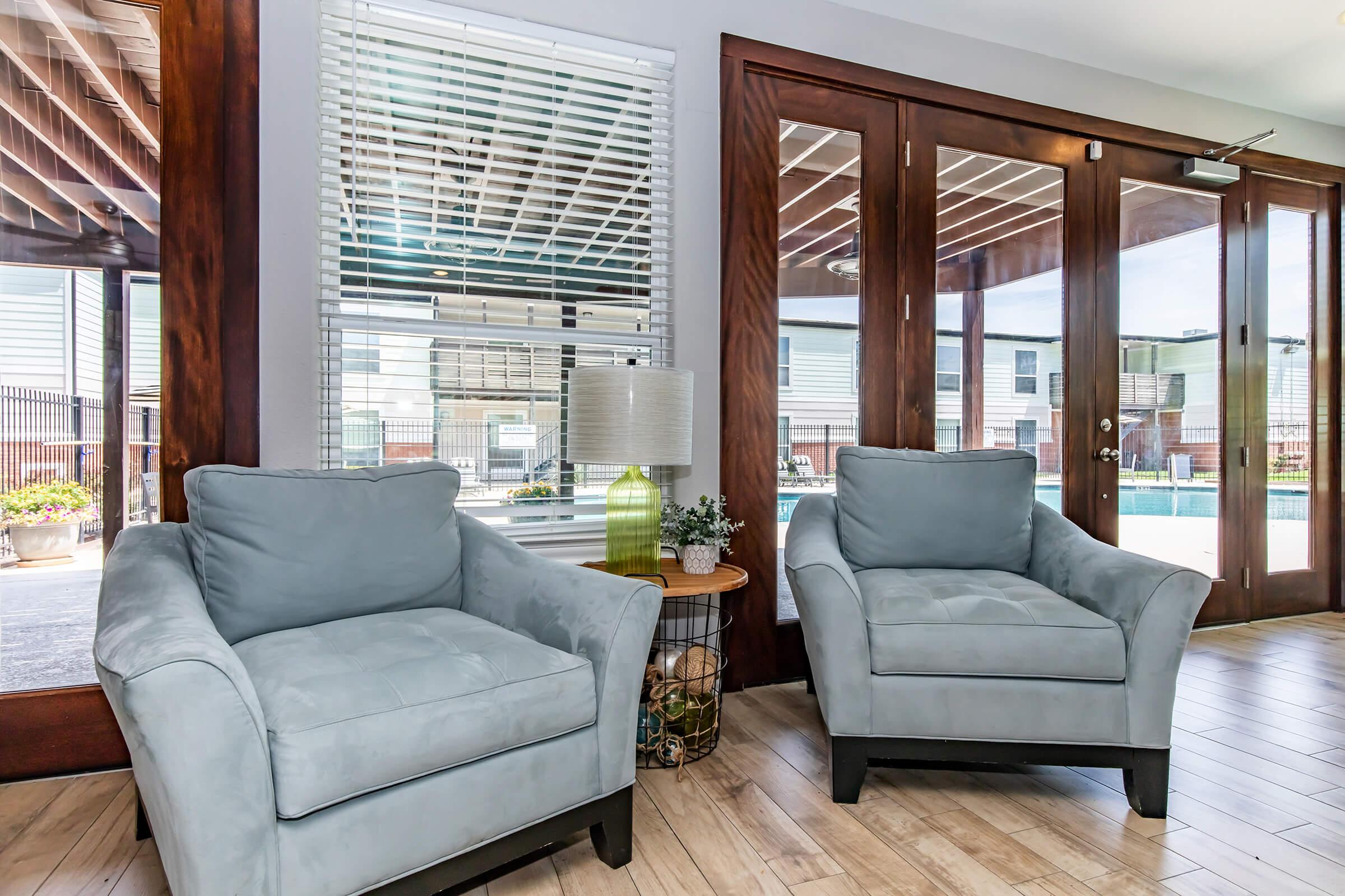 Two light blue armchairs are placed in a modern indoor space with wooden flooring. A small round table stands beside one chair, holding a lamp and decorative elements. Large windows allow natural light to enter, revealing a view of a pool area outside. The overall ambiance is bright and inviting.