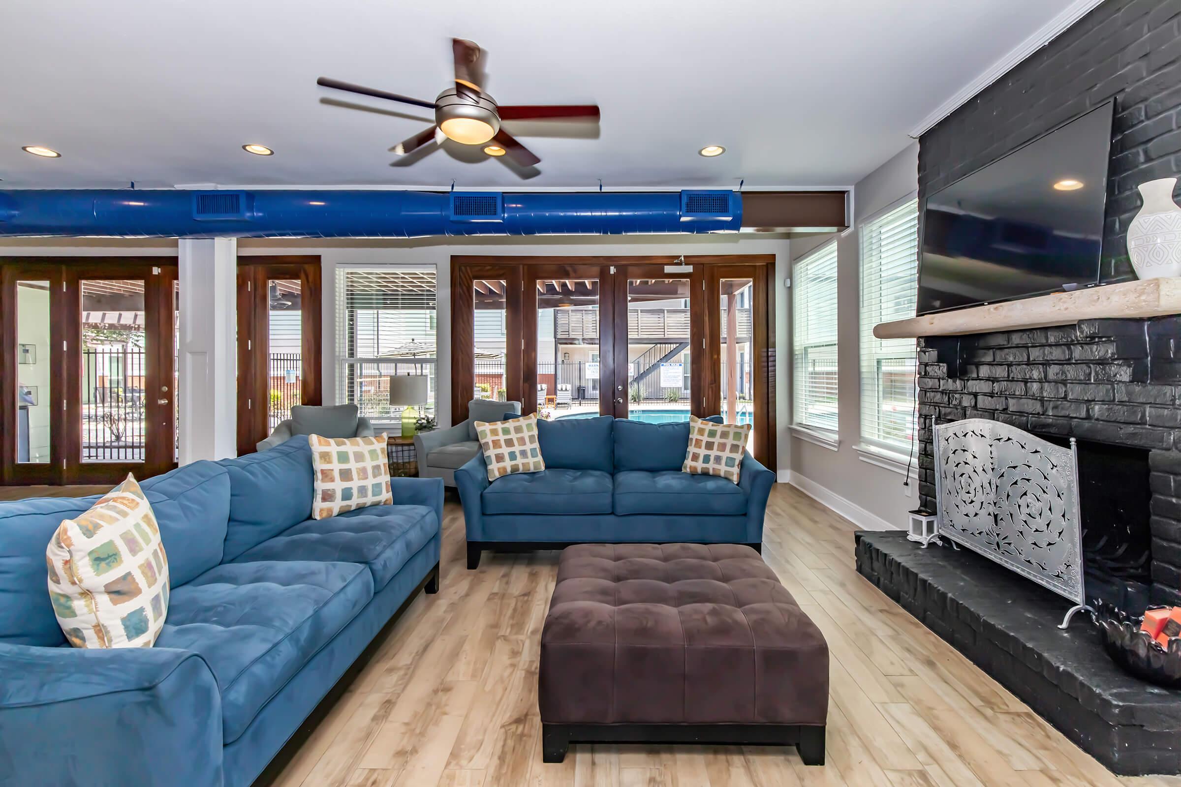 A modern living room featuring two blue sofas with decorative pillows, a dark brown ottoman in the center, and a sleek black fireplace. Large windows provide natural light, and a ceiling fan adds to the comfortable atmosphere. Decor includes a stylish TV mounted on the wall and a cozy, inviting ambiance.