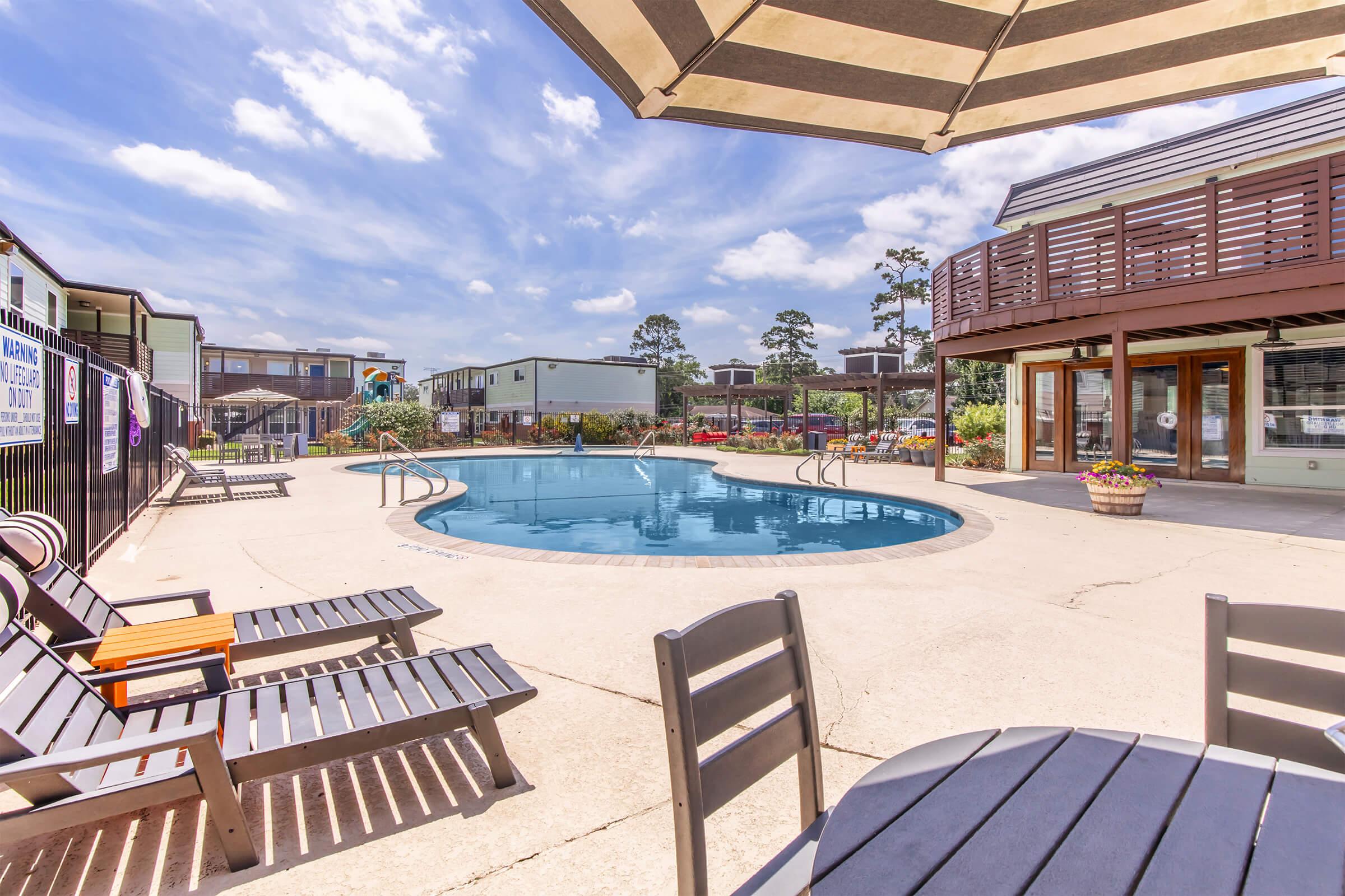 A spacious outdoor area featuring a sparkling blue swimming pool surrounded by lounge chairs and umbrellas. In the background, there are multiple buildings and gardens, with a clear blue sky overhead and a few fluffy clouds, creating a relaxed and inviting atmosphere.