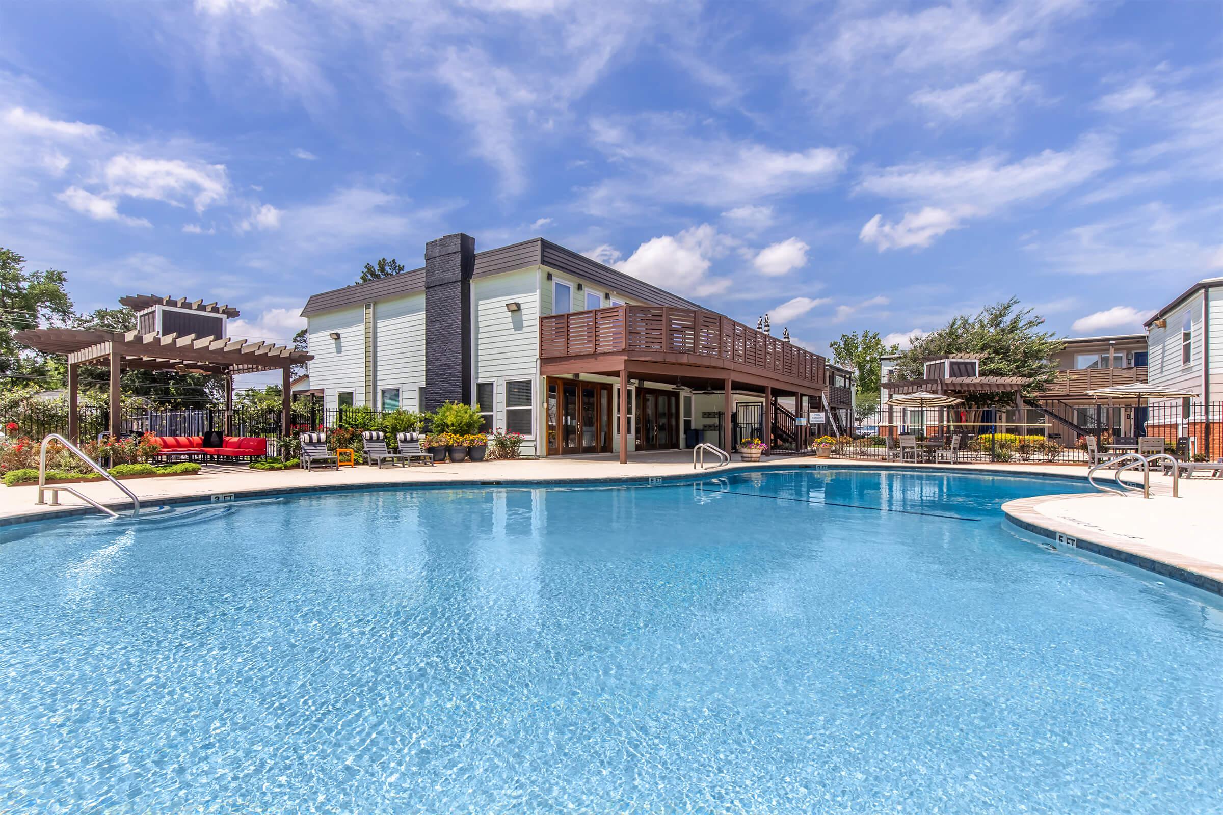 A clear swimming pool in the foreground with a wooden deck and lounge chairs. Beyond the pool, a residential building features large windows and a balcony. Colorful flowers and greenery surround the area, with a pergola structure on the left. The sky is partly cloudy, creating a bright and inviting atmosphere.