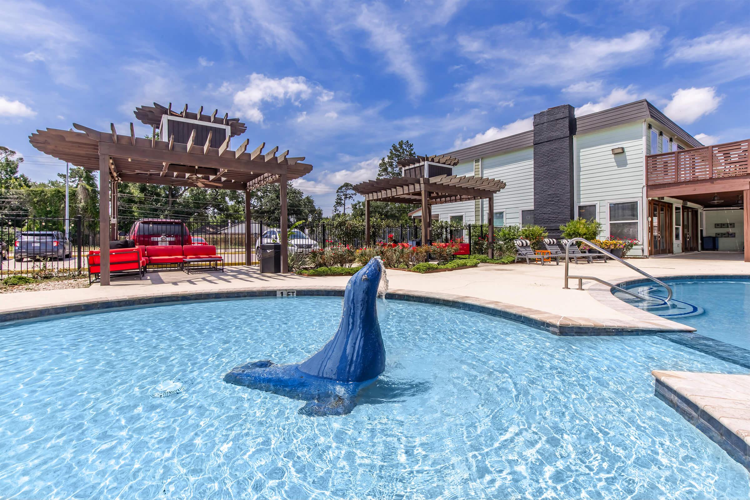 A resort-style swimming pool featuring a blue seal statue rising from the water. Surrounding the pool are lounge chairs, vibrant landscaping, and a pergola. A building with multiple stories is in the background under a clear blue sky with scattered clouds.