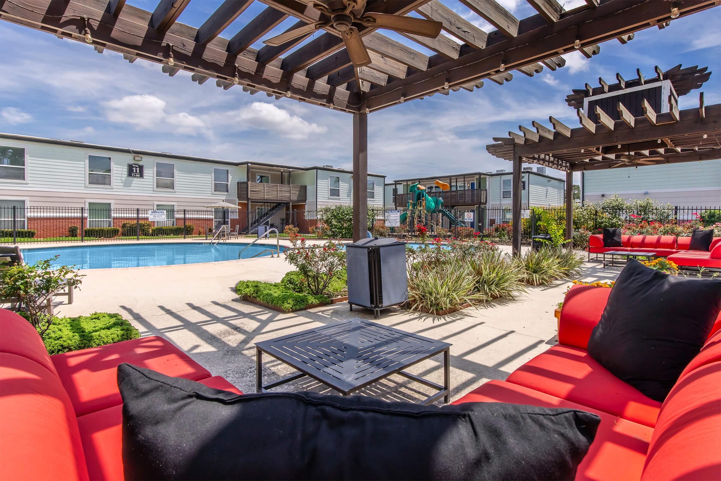 Outdoor view of a swimming pool surrounded by lounge seating with red cushions and a shaded pergola. Nearby greenery and a playground are visible in the background, along with two-story buildings. The sky is partly cloudy, enhancing the relaxed atmosphere.