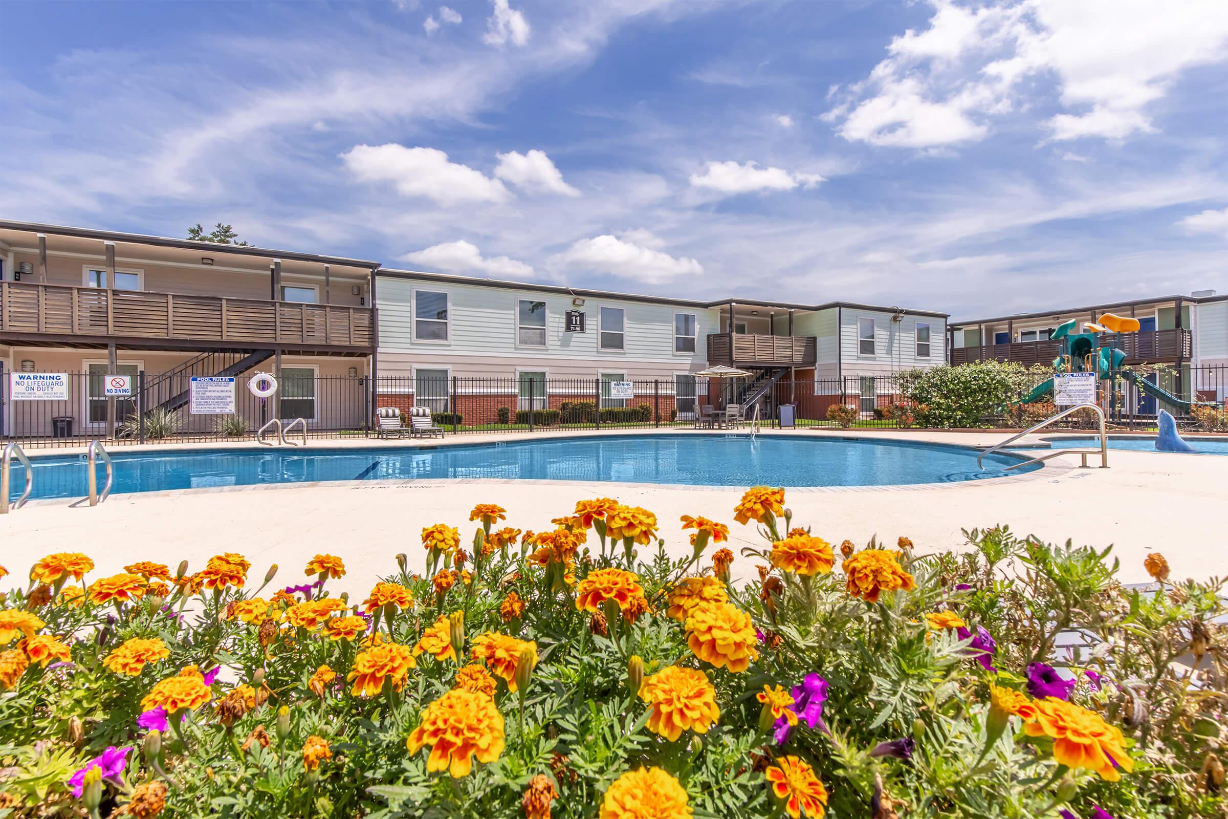 A clear swimming pool surrounded by vibrant flower beds featuring colorful marigolds. In the background, there are multi-story apartment buildings and a playground area, all under a bright blue sky with scattered clouds. The scene conveys a welcoming and relaxing atmosphere.