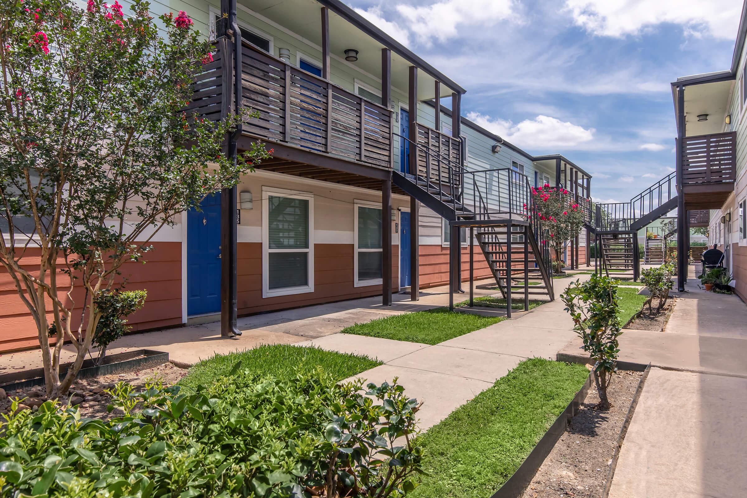 A sunny residential complex featuring multiple two-story buildings with balconies. They are surrounded by well-maintained grassy areas and bushes. A concrete walkway runs through the center, leading to each entrance, while a staircase is visible on the side of the buildings. The sky is bright blue with some clouds.