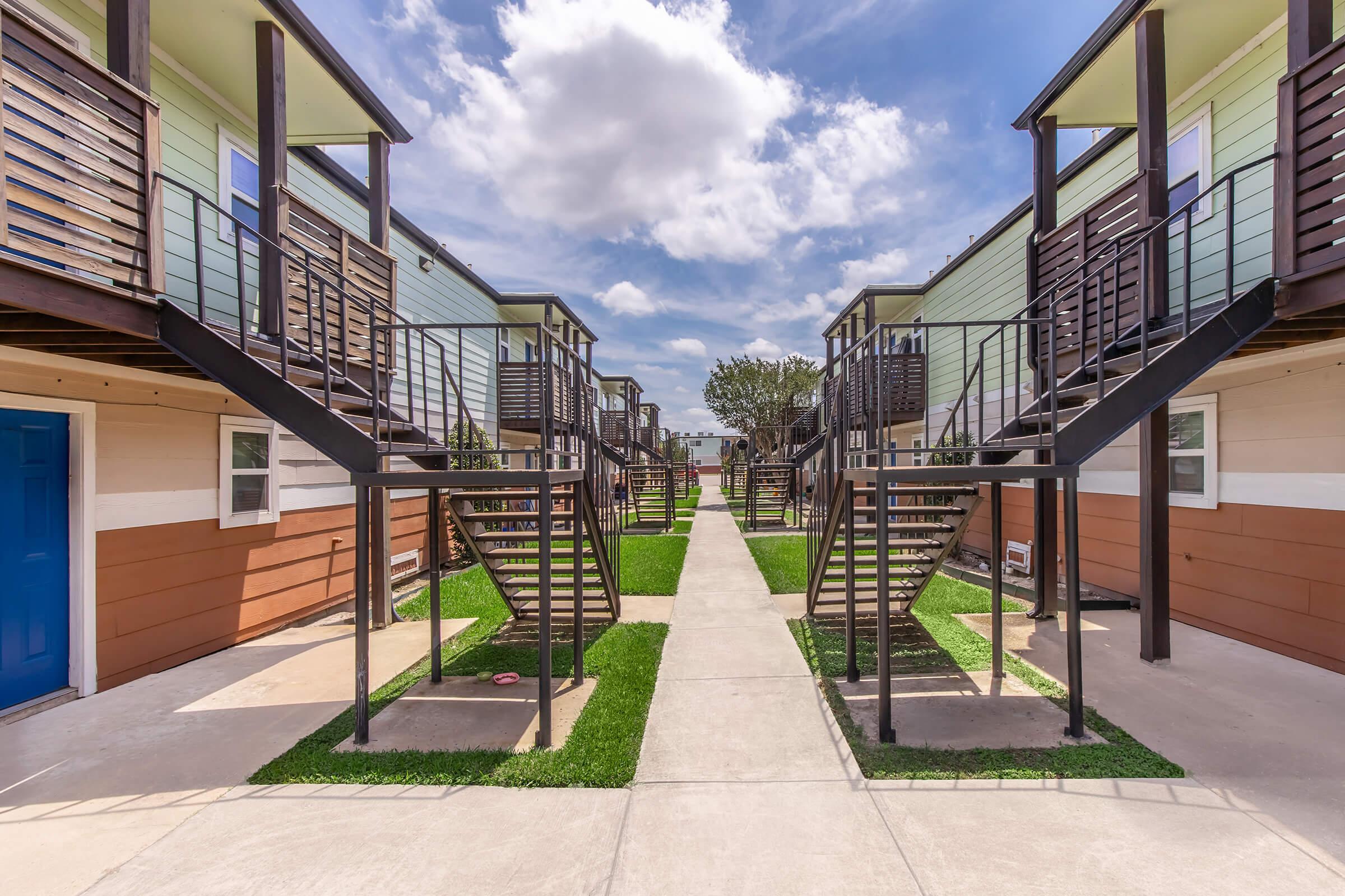 A view of a row of small, modern apartment buildings arranged in a U-shape, featuring green lawns and pathways. Each building has a staircase leading to an upper level, and the sky is partially cloudy, creating a bright atmosphere.
