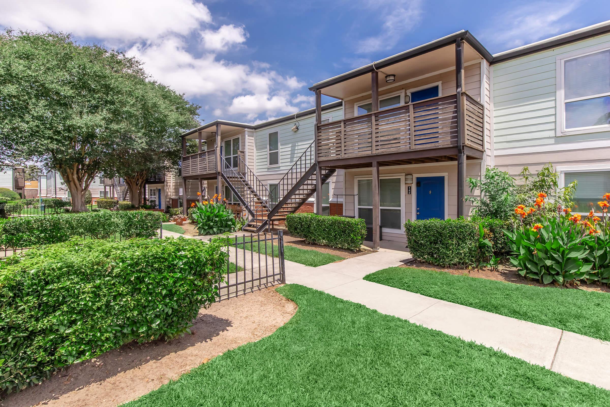 A well-kept apartment complex featuring two-story buildings with wooden balconies. Lush green lawns and neatly trimmed hedges surround the pathways. Colorful flowers add vibrancy to the landscape, and a gated entrance leads into the area under a partly cloudy sky.
