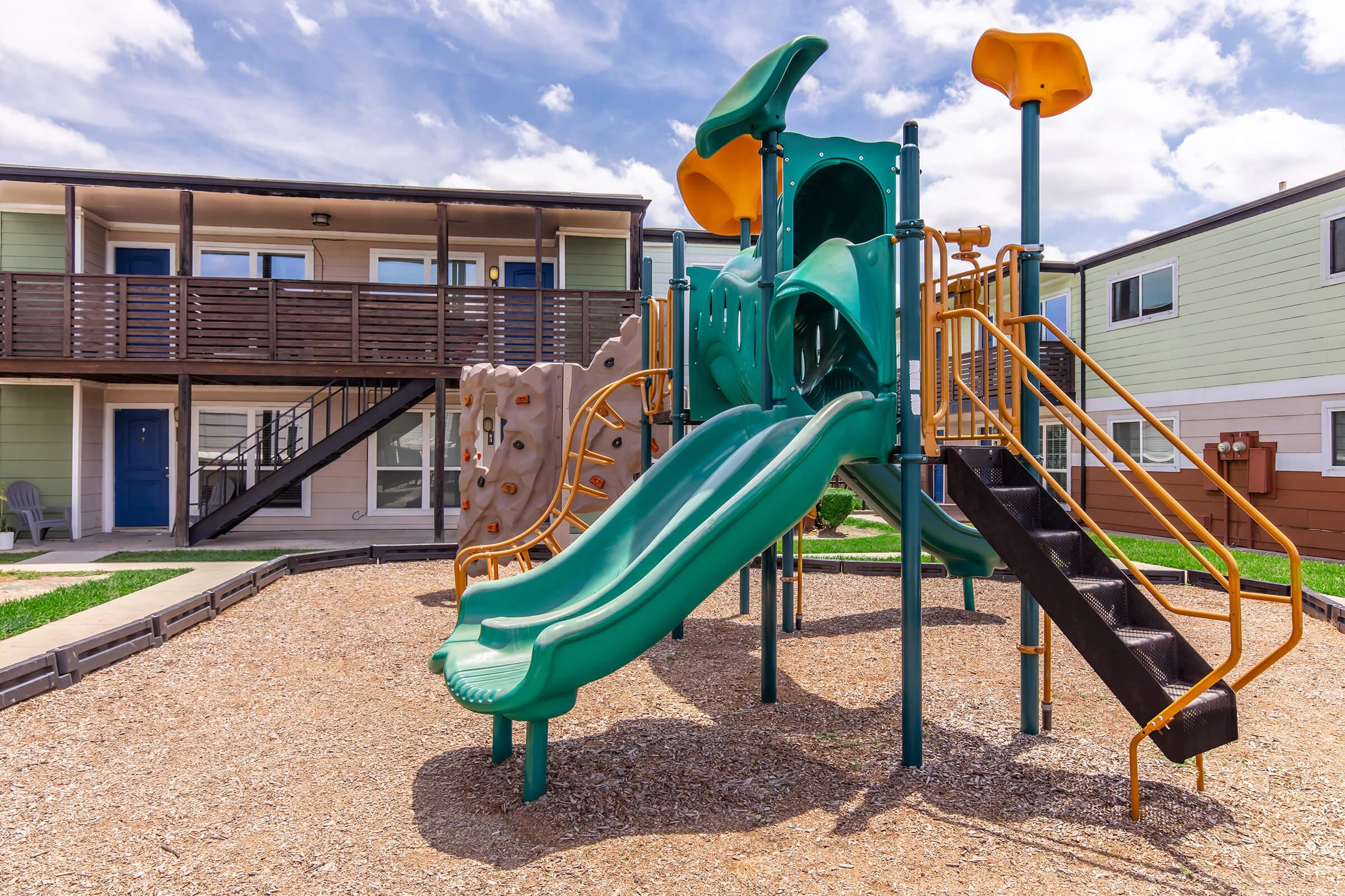 A colorful playground featuring two green slides, climbing structures, and jungle gym elements, set in a gravel area. In the background, there is a two-story apartment building with wooden balconies and various colored exterior walls. The sky is partly cloudy.