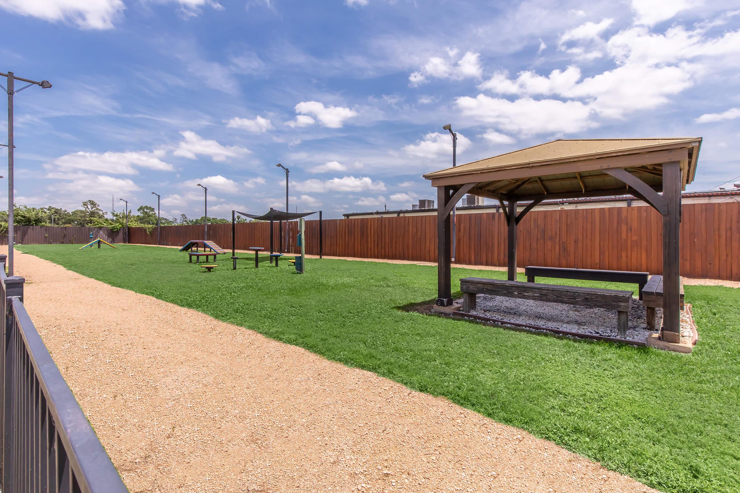 A sunny dog park featuring a spacious, grassy area with a shaded gazebo and benches. There are playground structures for dogs, such as agility equipment, along a gravel path and surrounded by wooden fences. The sky is partly cloudy, creating a bright and inviting atmosphere.