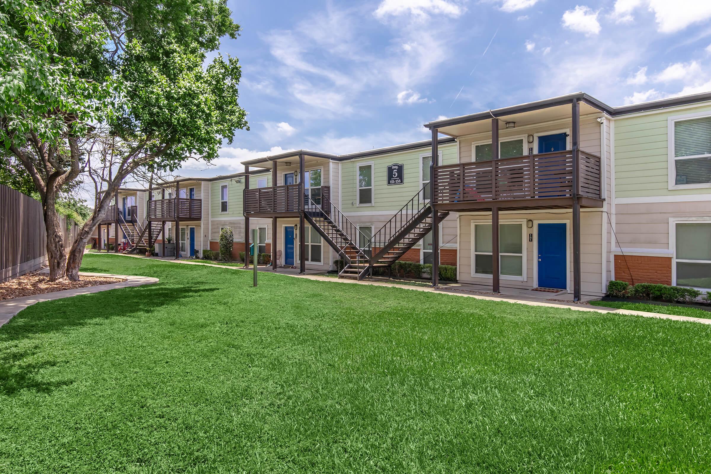 A view of a residential complex featuring two-story apartment buildings with balconies, surrounded by lush green grass and trees. The buildings have a light color scheme with blue doors. A pathway leads through the well-maintained lawn, showcasing a pleasant outdoor living space.