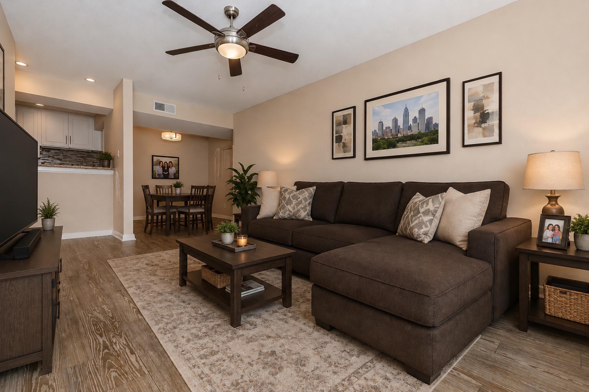 A cozy living room featuring a dark brown sectional sofa with decorative pillows, a wooden coffee table, and a light-colored rug. There is a flat-screen TV on a TV stand, a plant in the corner, and framed pictures on the walls. In the background, a dining area with a table and chairs is visible. Ceiling fan above.