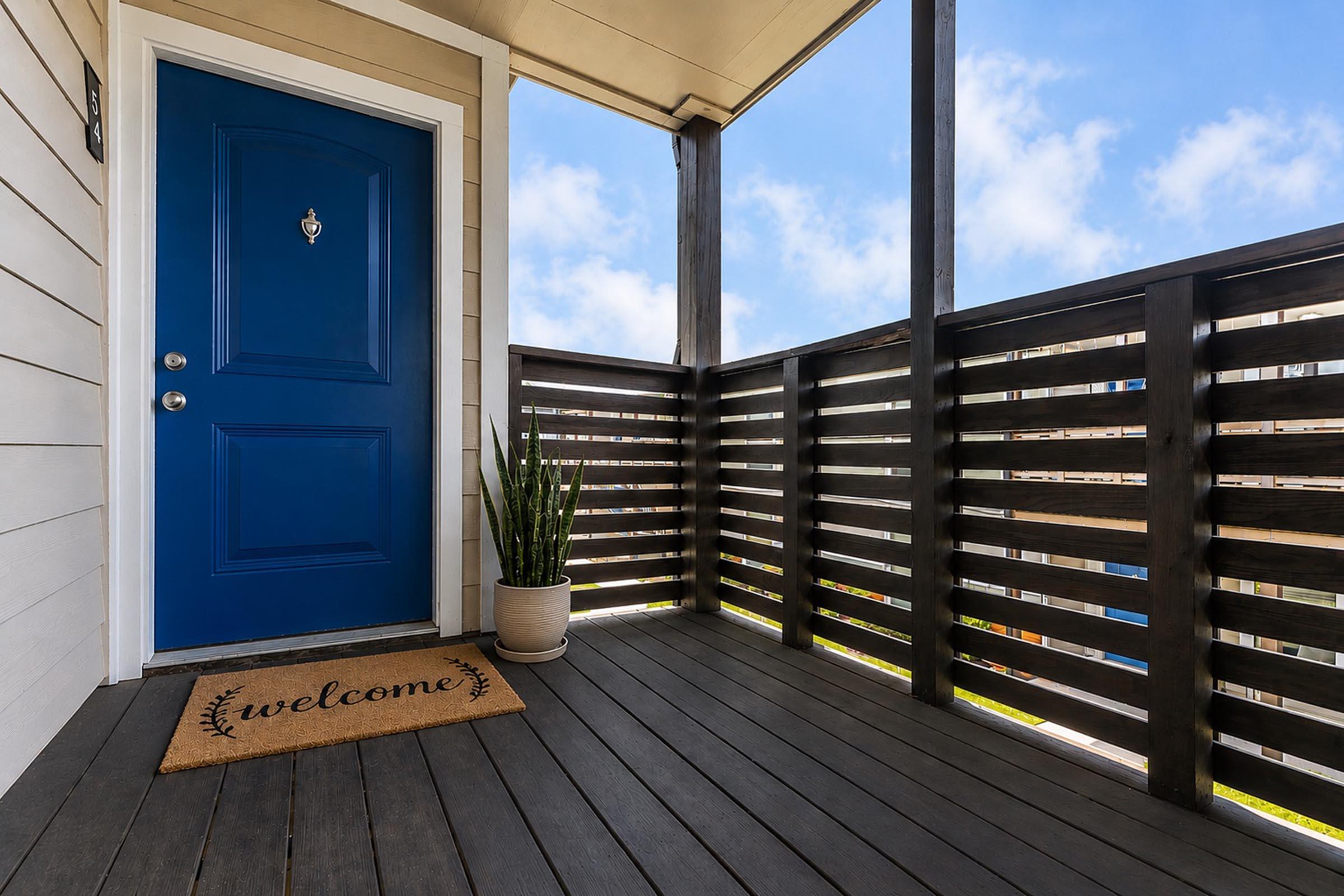 A bright blue front door set against a light-colored wall, with a welcome mat at the entrance. There’s a modern planter with a tall green plant beside the door. The porch features a wooden railing with horizontal slats and a sunny sky in the background.