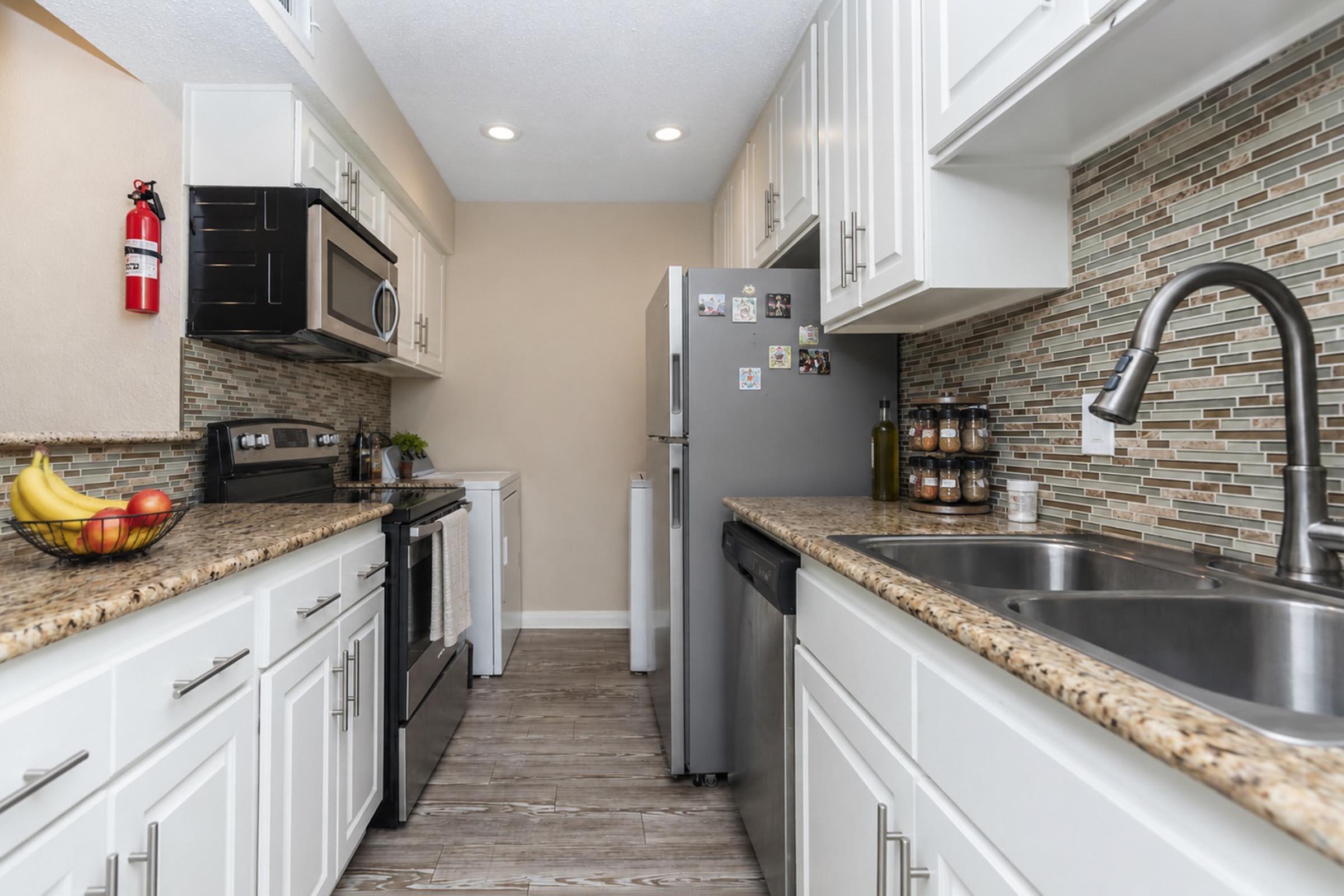 A modern kitchen featuring white cabinets, a granite countertop, stainless steel appliances including a microwave and refrigerator, and a dual sink. The backsplash has a mosaic design, and there are decorative elements like a fruit bowl and various items on the countertop. The space is well-lit and organized.