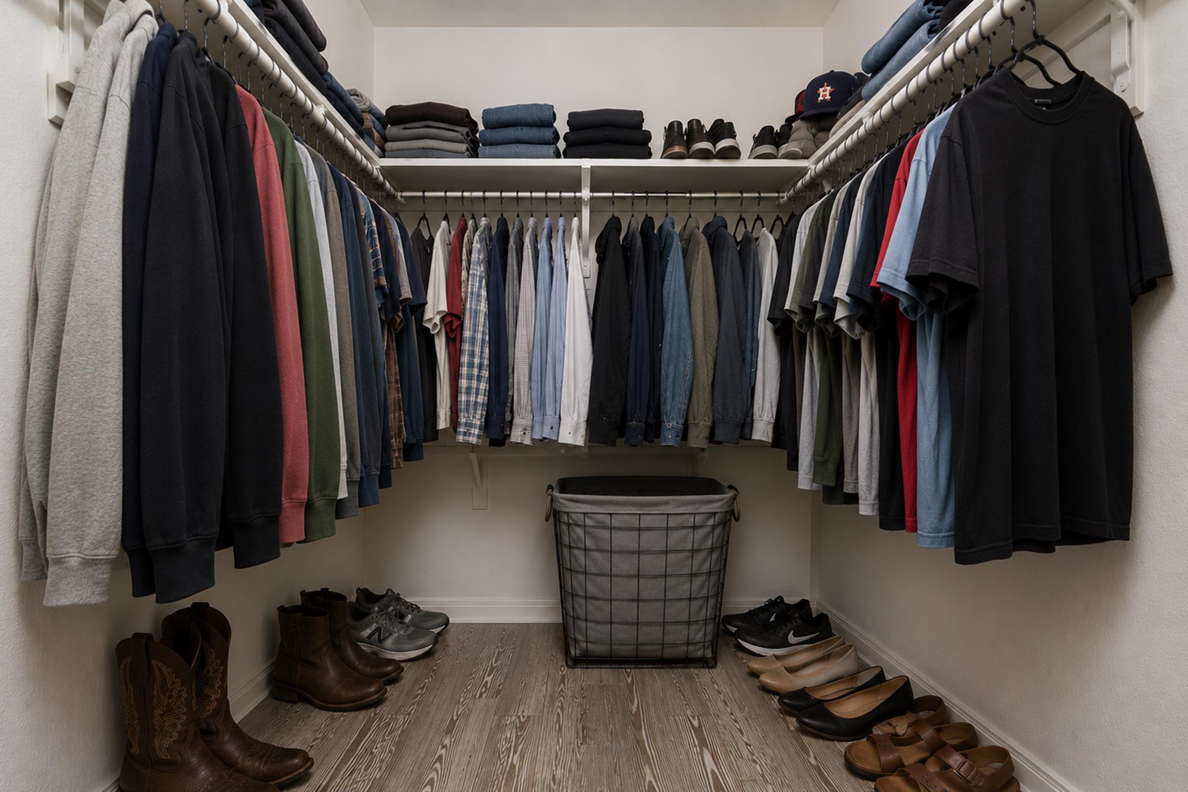 A well-organized closet featuring two rows of hanging clothes including shirts and sweatshirts in various colors. On the floor, there are several pairs of shoes, including boots and sandals, and a laundry basket. The walls are painted white, providing a clean and bright appearance to the space.