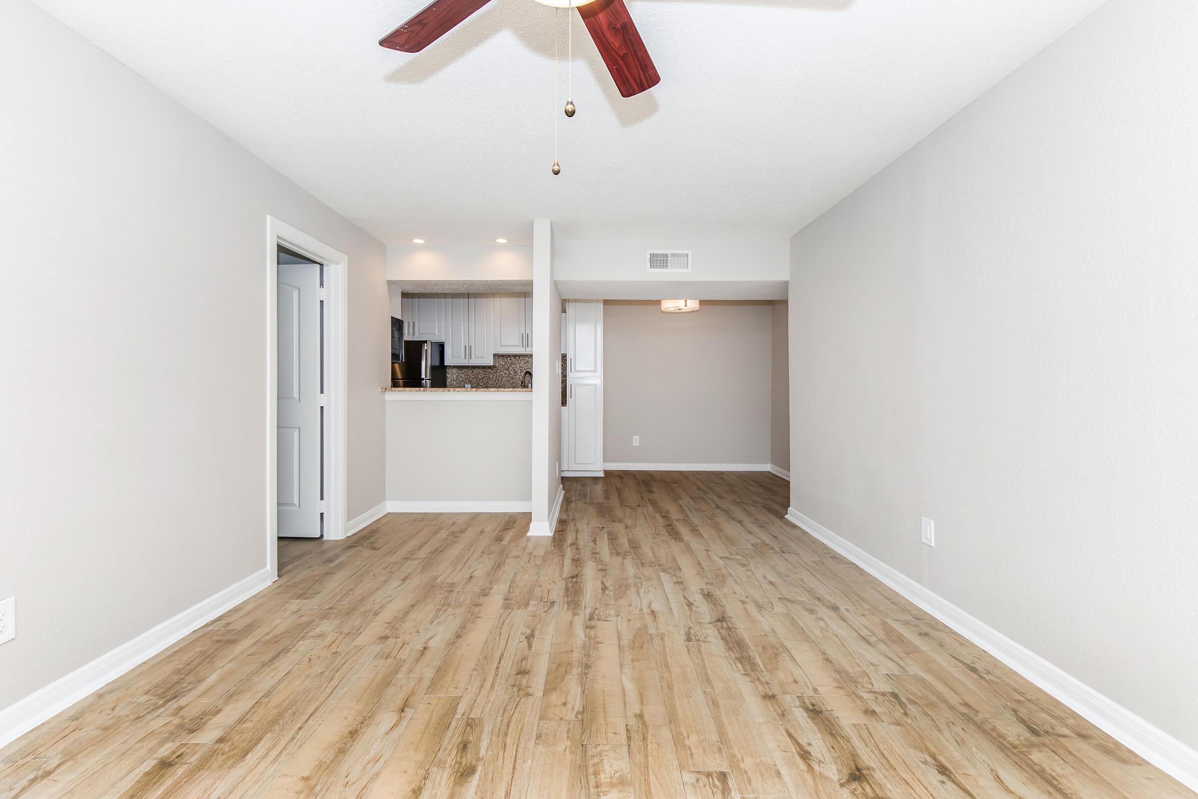 A spacious, well-lit living area featuring light wood flooring, a ceiling fan, and neutral-colored walls. An open kitchen is visible in the background, with modern cabinetry and appliances. The layout creates an inviting atmosphere, ideal for relaxation or entertaining.