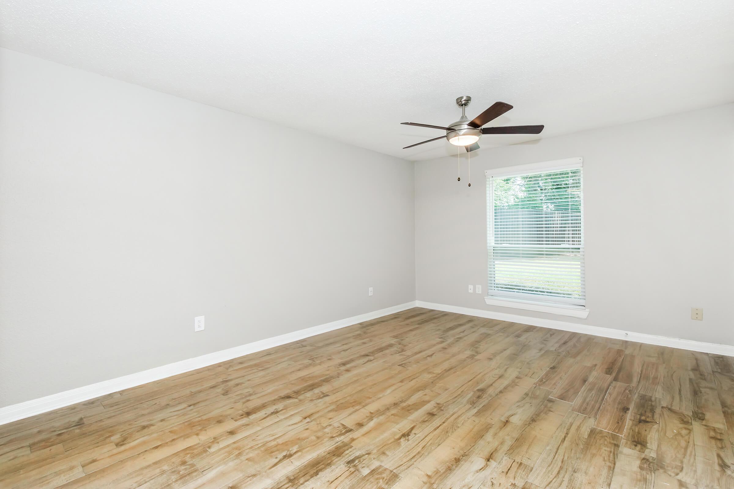 A spacious, empty room with light beige walls and a ceiling fan. The floor is covered in wooden laminate, and there is a window with blinds allowing natural light to enter. The overall appearance is clean and modern, providing a blank canvas for furnishing and decor.