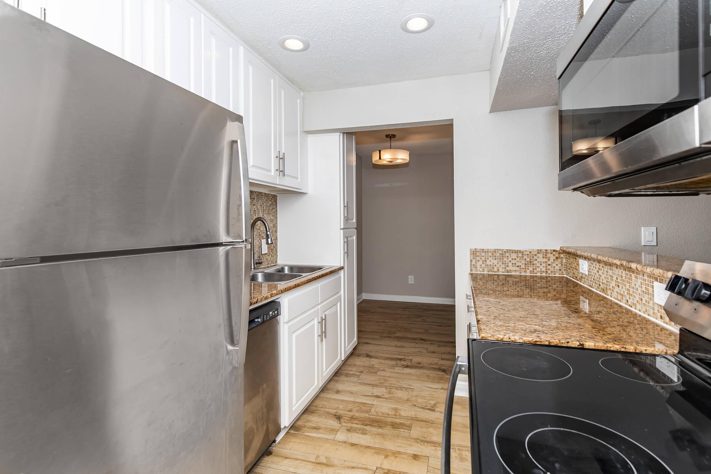 A modern kitchen featuring stainless steel appliances, including a refrigerator and oven, along with a dishwasher. The countertops are made of granite, complemented by white cabinetry. The kitchen has wooden flooring and a light fixture visible in the adjoining area. The overall design is contemporary and well-lit.