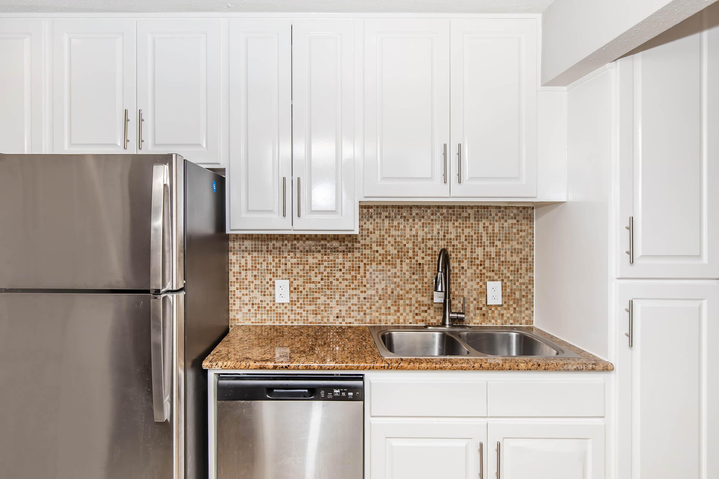 Modern kitchen featuring stainless steel appliances, including a refrigerator, dishwasher, and a double sink. The countertops are made of granite, and the backsplash consists of small, textured tiles in earthy tones. White cabinetry provides a clean, bright aesthetic in the space.