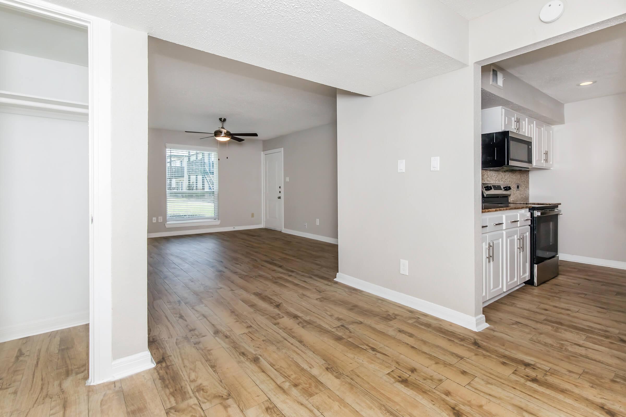 Spacious interior of a modern apartment featuring a living area with a ceiling fan, light-colored walls, and a large window. The kitchen is visible on the right with stainless steel appliances, white cabinets, and a countertop, while the left side shows a closet space. The flooring is light-colored wood.
