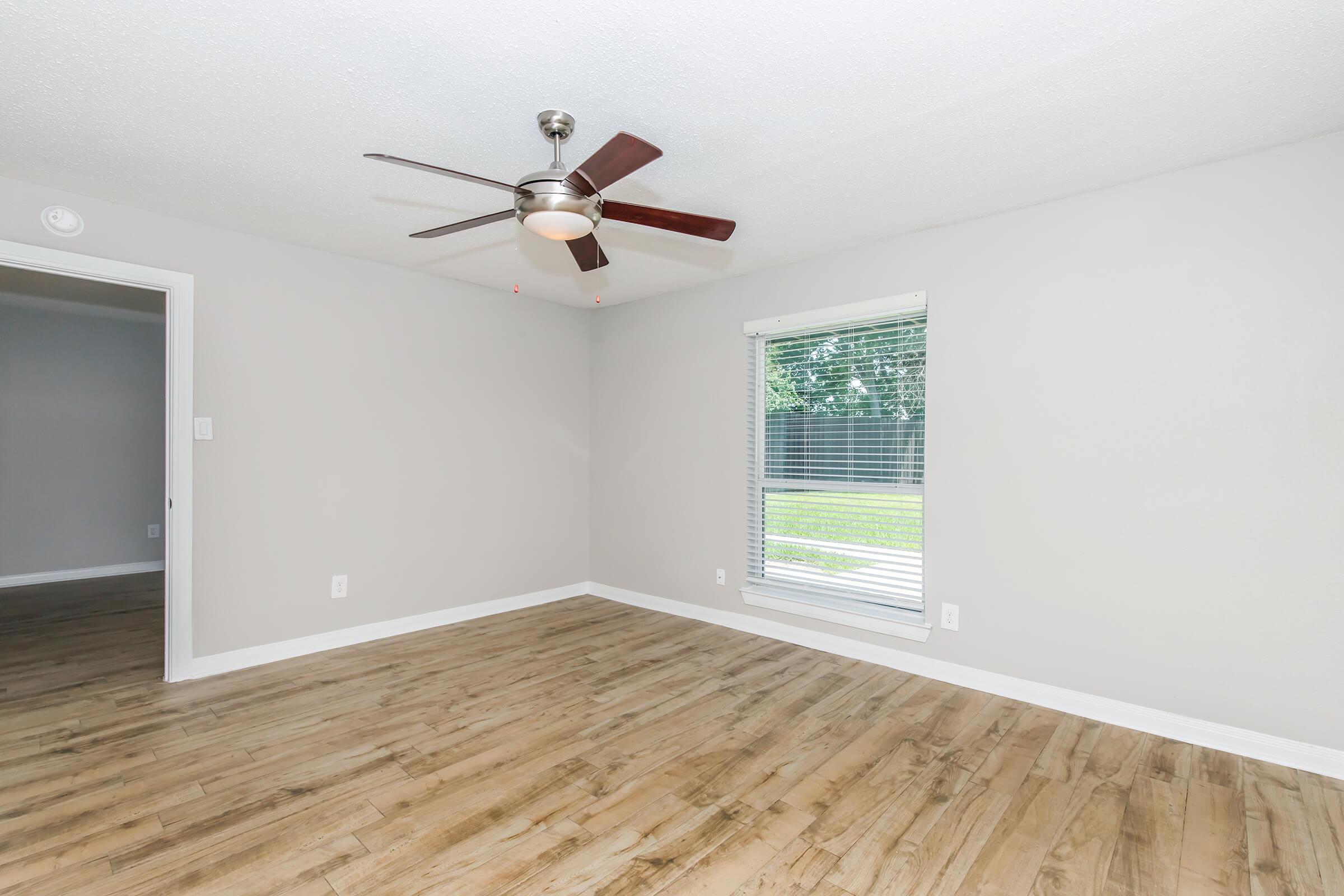 A well-lit, empty room featuring light-colored walls and a ceiling fan. The floor is made of wood, and there is a window with blinds allowing natural light in, overlooking a grassy area outside. A doorway is visible on the left leading to another space.
