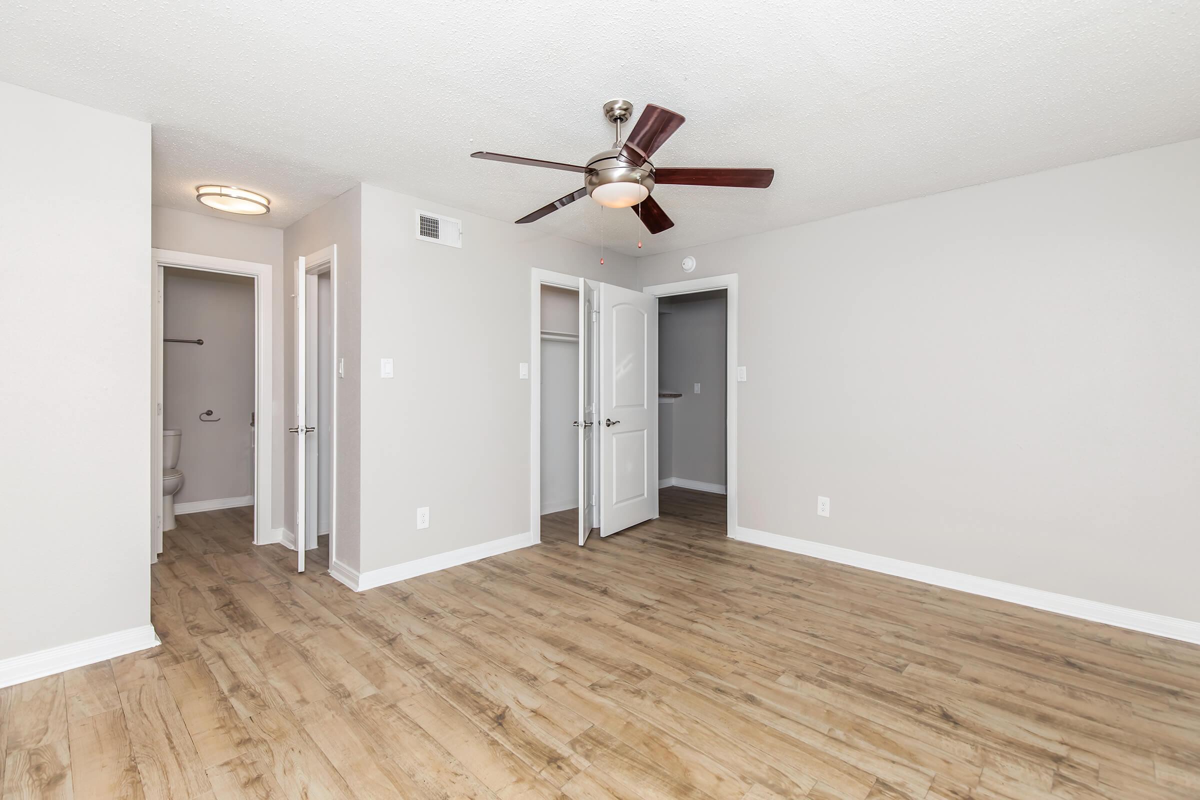 A spacious, empty room with light-colored walls and hardwood flooring. A ceiling fan is installed, and there are doors leading to other areas, including a bathroom. The natural light enhances the open feel of the space, making it bright and inviting.