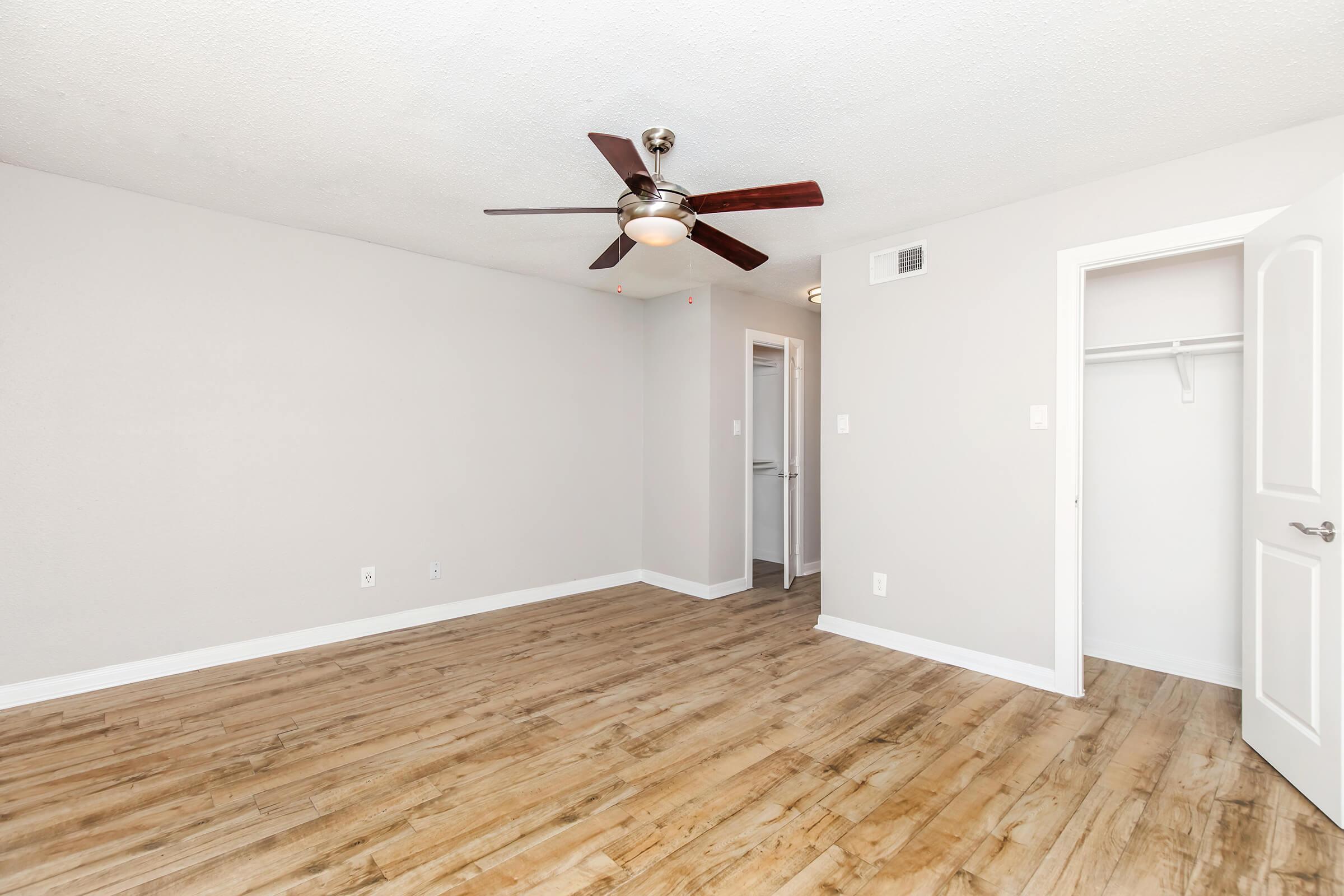 A spacious, empty room featuring light-colored walls and wooden flooring. A ceiling fan is mounted in the center, and there is a closet to the right with a white door. The room has a neutral and modern aesthetic, with natural light enhancing the open atmosphere.