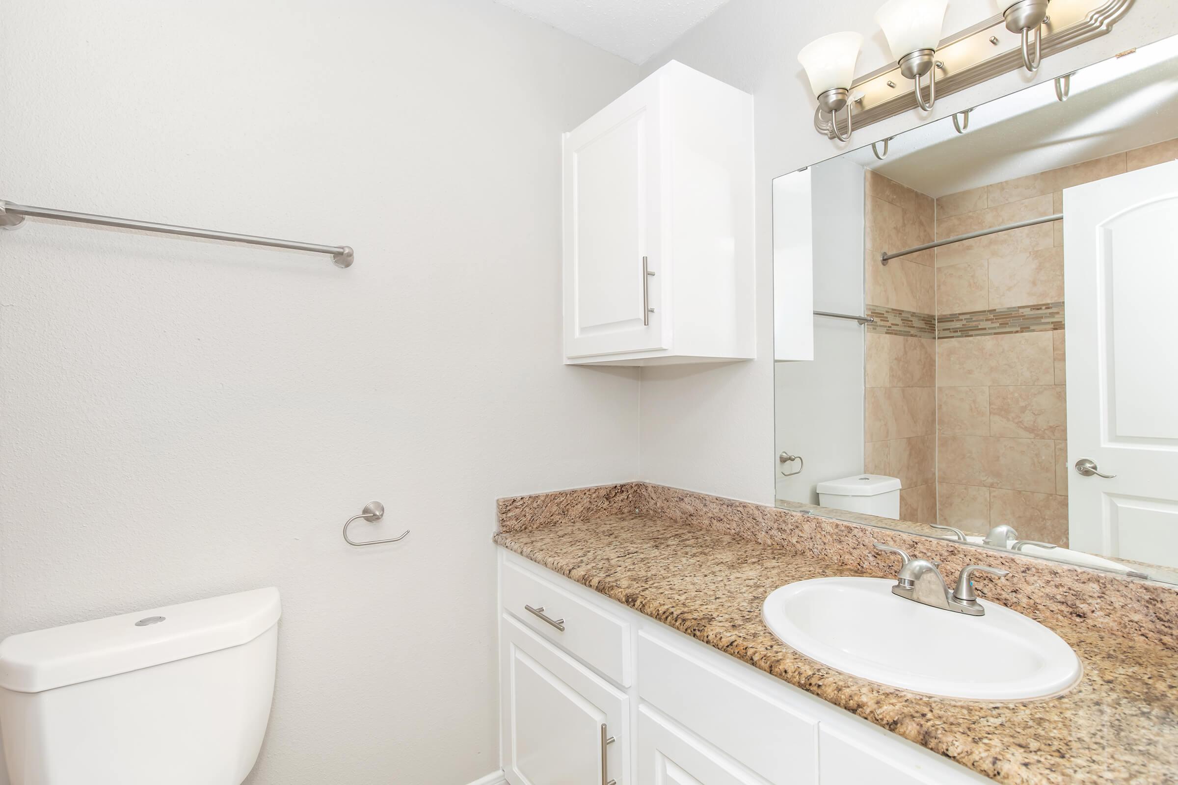 A clean and modern bathroom featuring a white toilet, a light-colored countertop with a sink, a mirror above the sink, and white cabinetry. Natural light fills the space, highlighting beige and tan tile accents on the walls and shower. A towel bar is mounted on the wall.