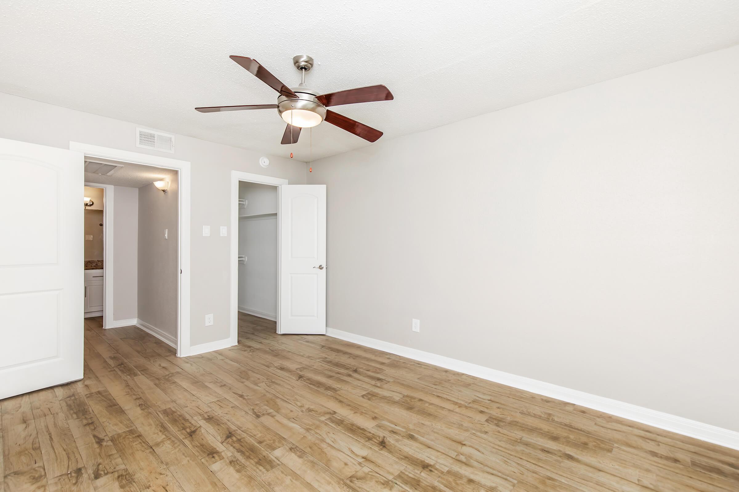 A well-lit, empty room featuring light-colored walls and a ceiling fan. There is a door leading to a closet and another door in the background that appears to lead to a bathroom. The floor is a warm, wooden finish, creating a spacious and inviting atmosphere.