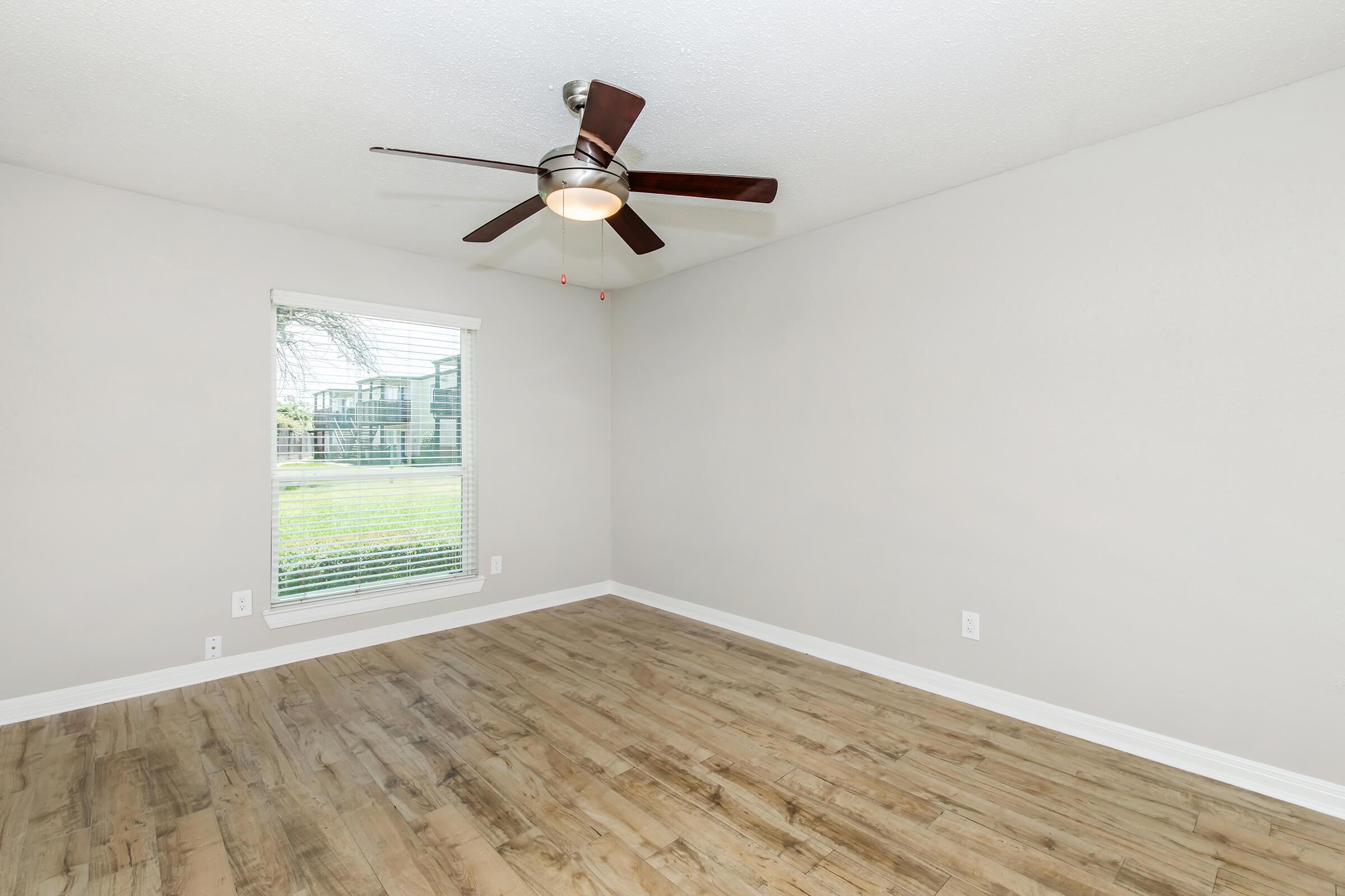 A spacious, empty room with light gray walls and laminate flooring. A ceiling fan with wooden blades spins above. A window with blinds lets in natural light, revealing a view of greenery outside. The room is unfurnished, creating a clean and simple aesthetic.