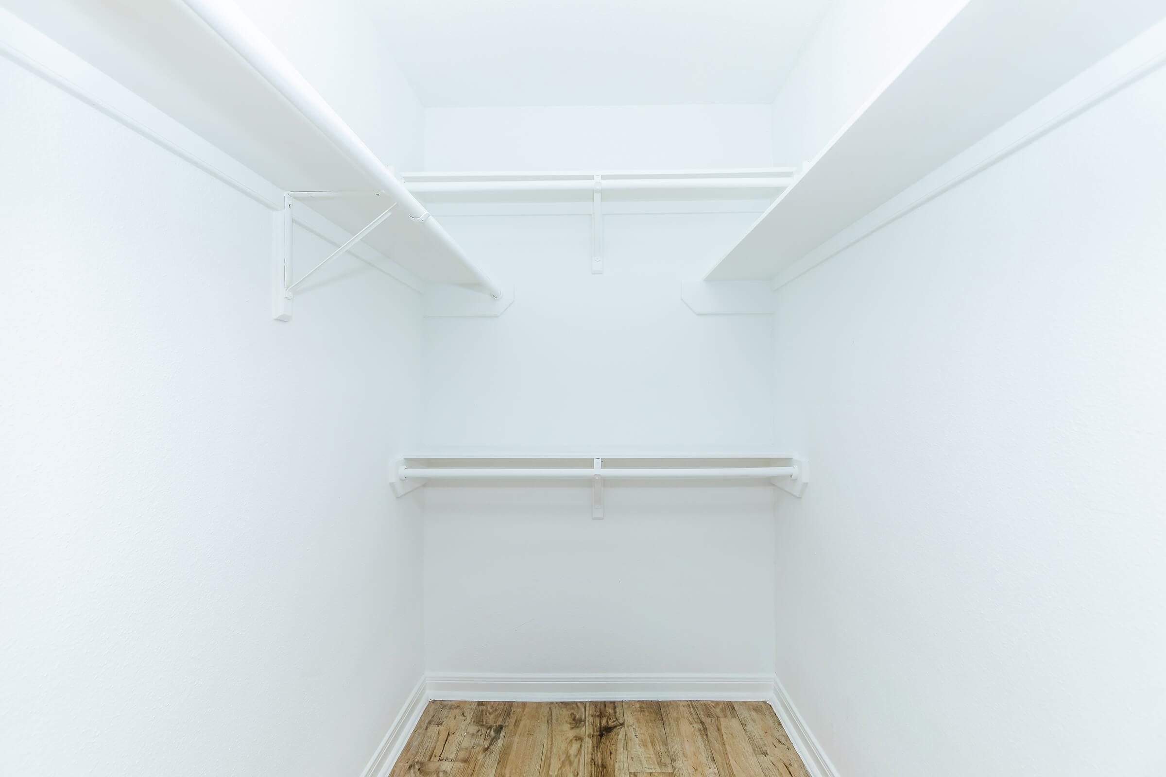A clean, empty closet with white walls and shelves. The closet features a light wooden floor and is well-lit, providing a minimalist and spacious appearance.