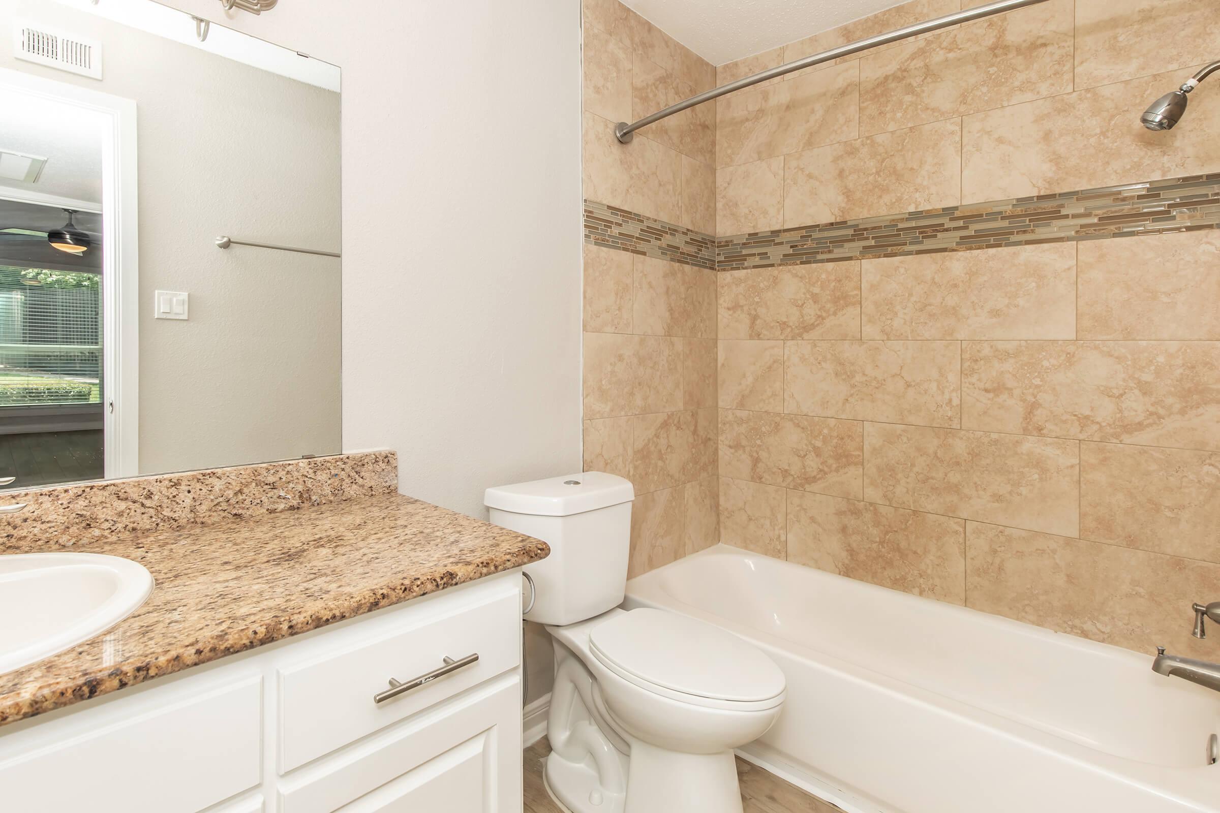 A modern bathroom featuring beige tiled walls, a white bathtub, and a toilet. The countertop is granite with a sink, and there is a showerhead above the bathtub. Natural light comes through a window, and a mirror hangs above the sink. The overall design is clean and contemporary.