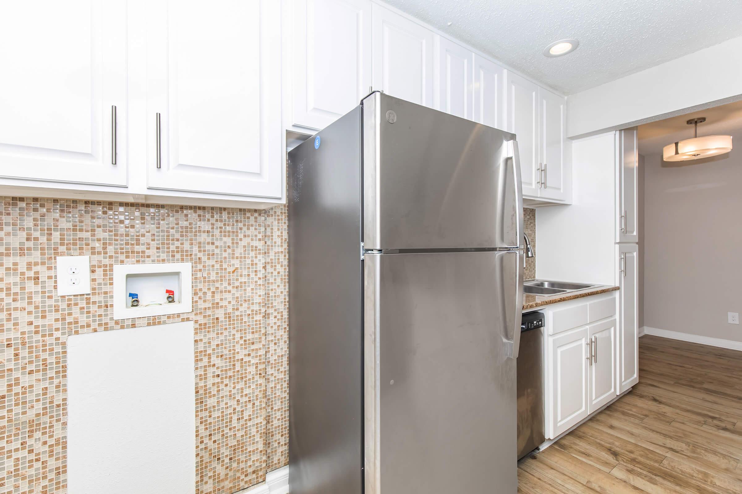 A modern kitchen featuring a stainless steel refrigerator, white cabinetry, and a tiled mosaic backsplash. The space includes a stainless steel sink and dishwasher, along with light wood flooring that adds warmth to the area. Bright lighting fixtures complete the contemporary design.