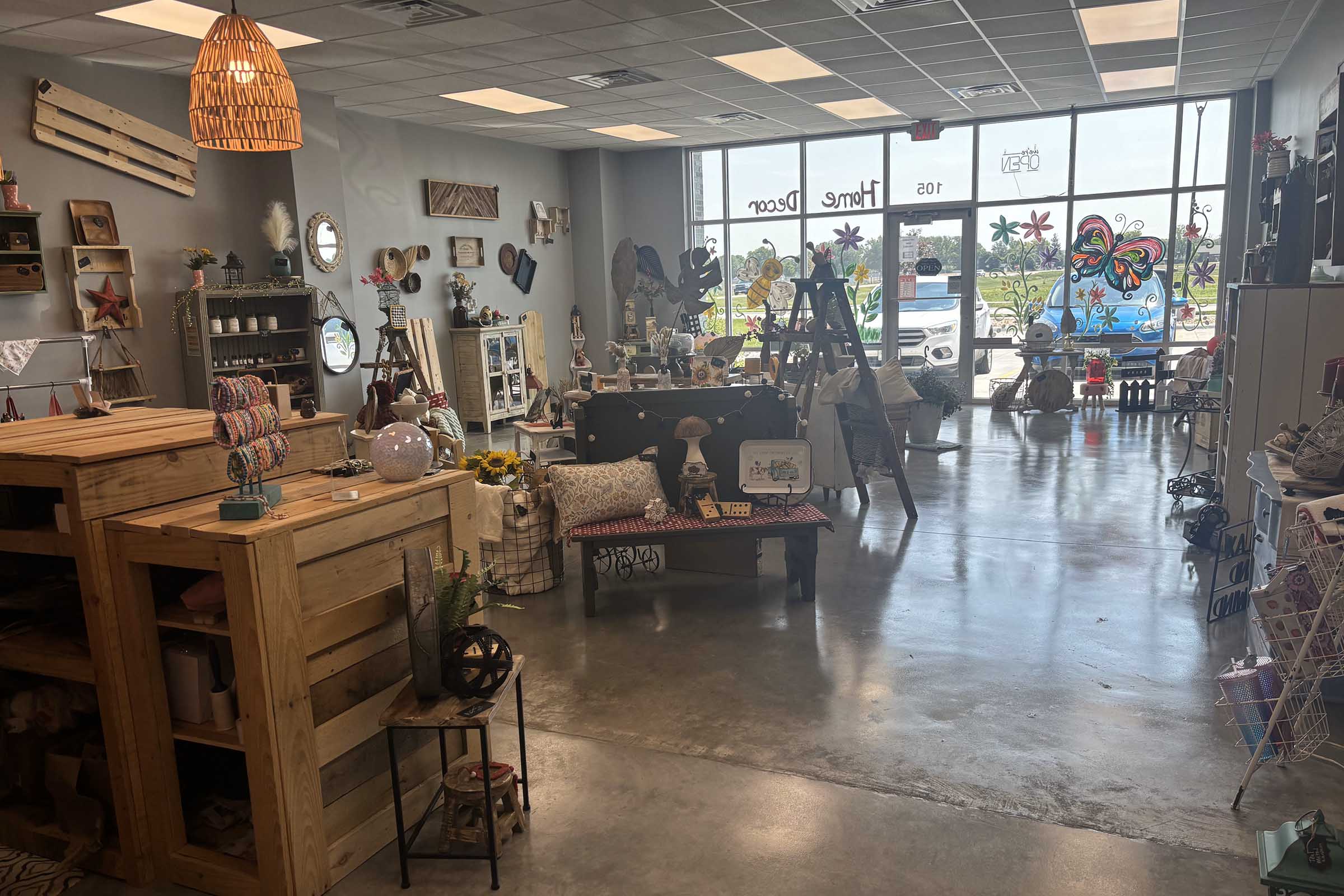 A cozy home decor shop featuring a variety of items including wooden furniture, decorative mirrors, and whimsical art pieces. The space is bright with large windows showcasing a colorful mural. Items like lamps, plants, and textiles are arranged to create an inviting atmosphere.