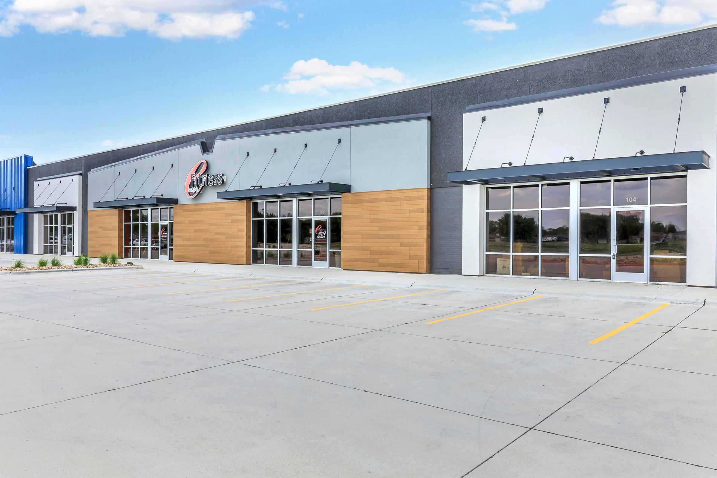 A modern commercial building with glass storefronts, featuring large windows and a neutral color palette. The area is landscaped with small plants, and the parking lot in front has several empty spaces. The sky is clear with a few clouds, creating a bright atmosphere.