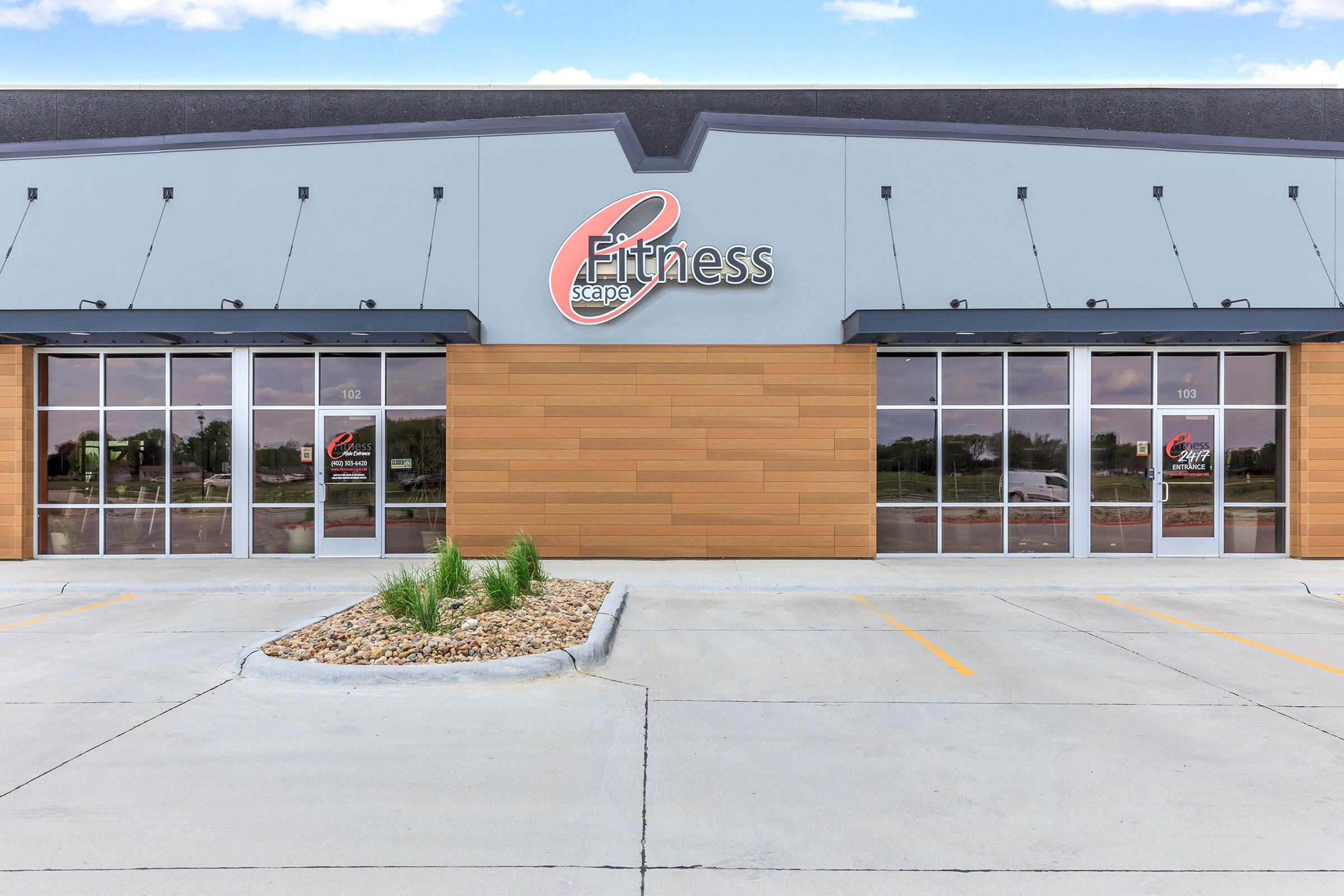 Exterior view of a Fitness Scope building, featuring a modern design with large glass windows and a contrasting wooden facade. The logo is prominently displayed above the entrance. A small landscaped area with rocks and grass is visible in front of the entrance, and the parking lot is seen in the foreground.